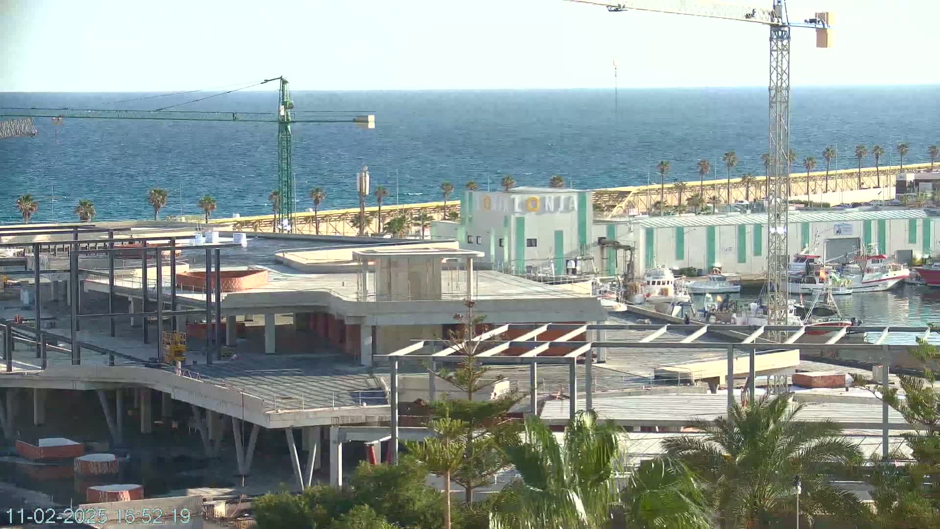 A partially constructed building overlooks a harbor filled with fishing boats under a clear sky and calm sea.