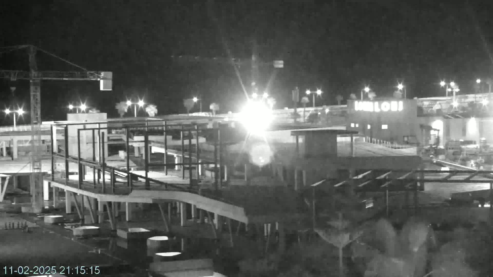 A nighttime, black and white image shows a building under construction next to a brightly lit structure, with a construction crane in the background.