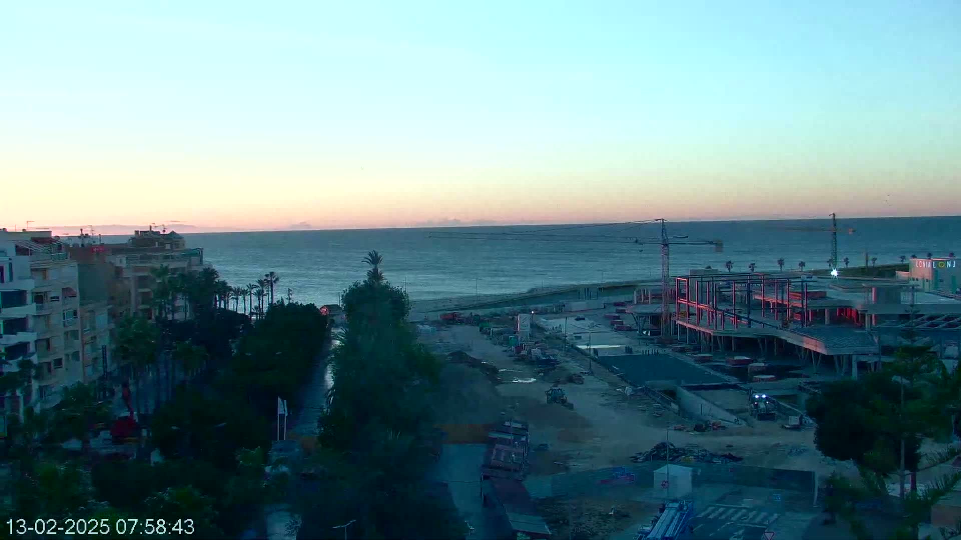 A sunrise over a calm sea, showing a coastal city with buildings and a large construction site with cranes.