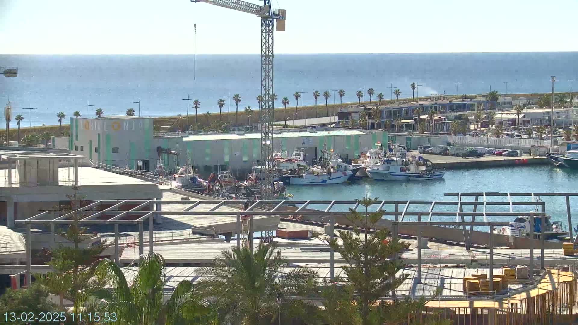 A harbor filled with fishing boats under a sunny sky, with a partially constructed building and a construction crane in the foreground.
