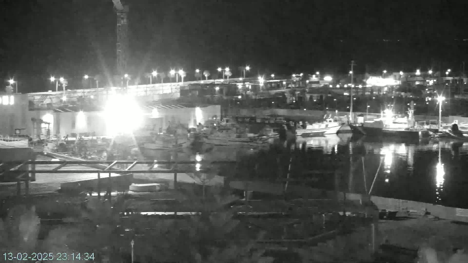 A nighttime, brightly lit harbor scene shows numerous boats docked at a pier with construction visible on the shore.
