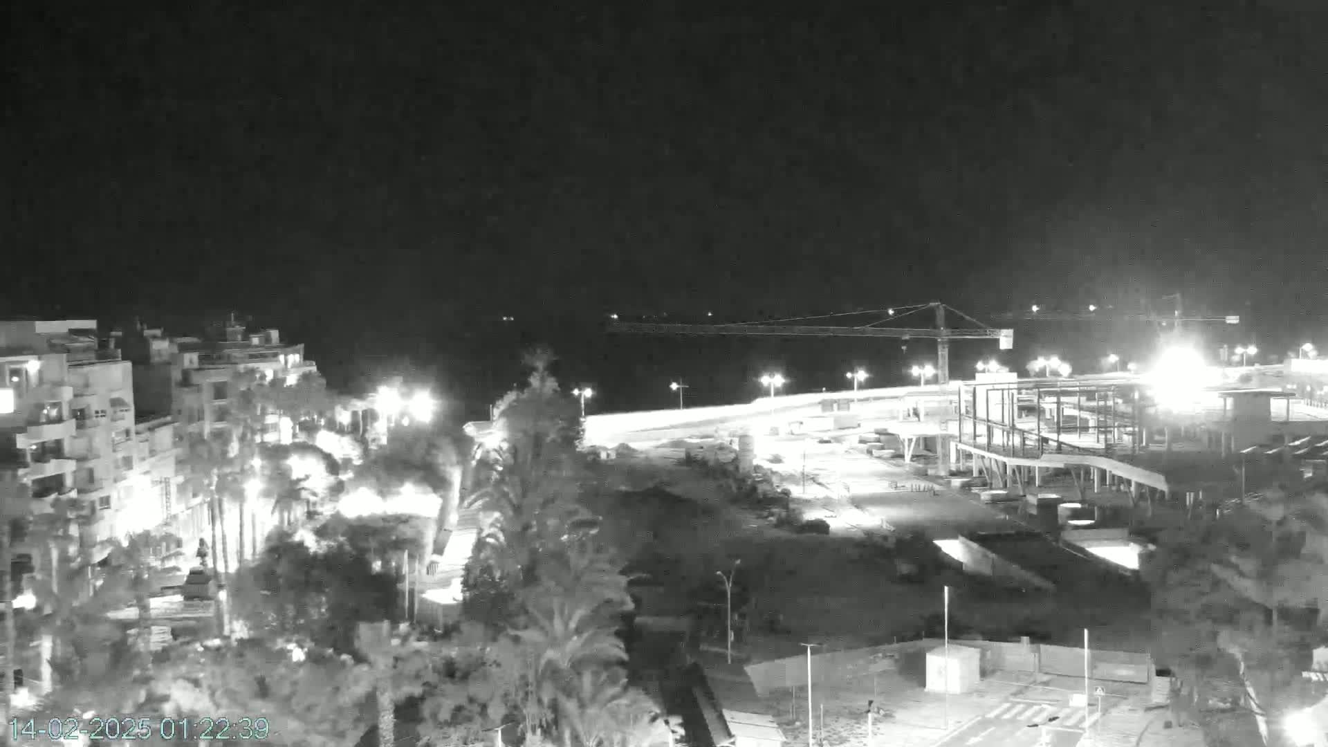 A nighttime, high-angle view shows a coastal town with buildings, palm trees, and a construction site under a clear, dark sky.