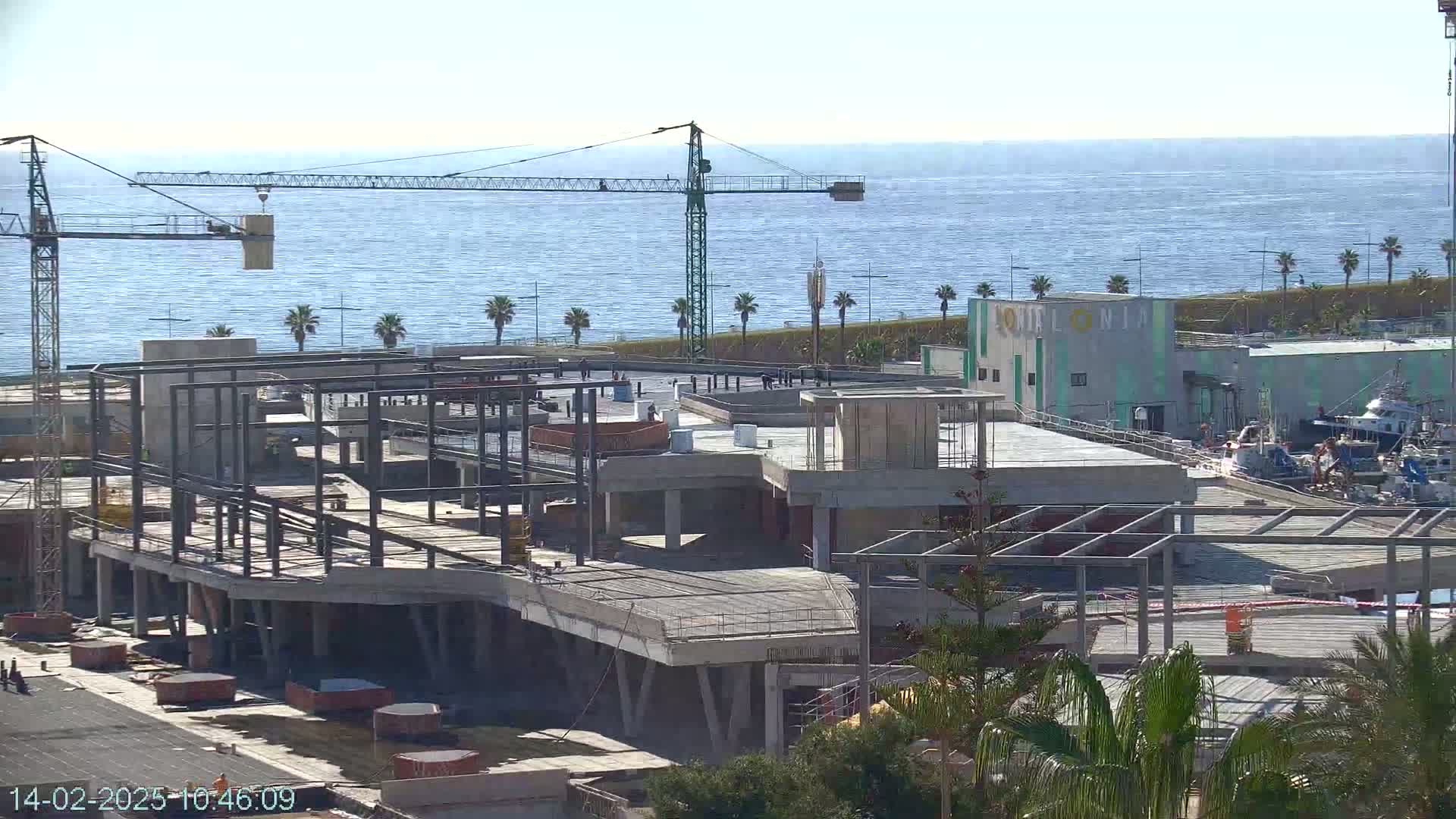 A large building under construction is situated near a calm ocean, with a construction crane visible against a clear sky.