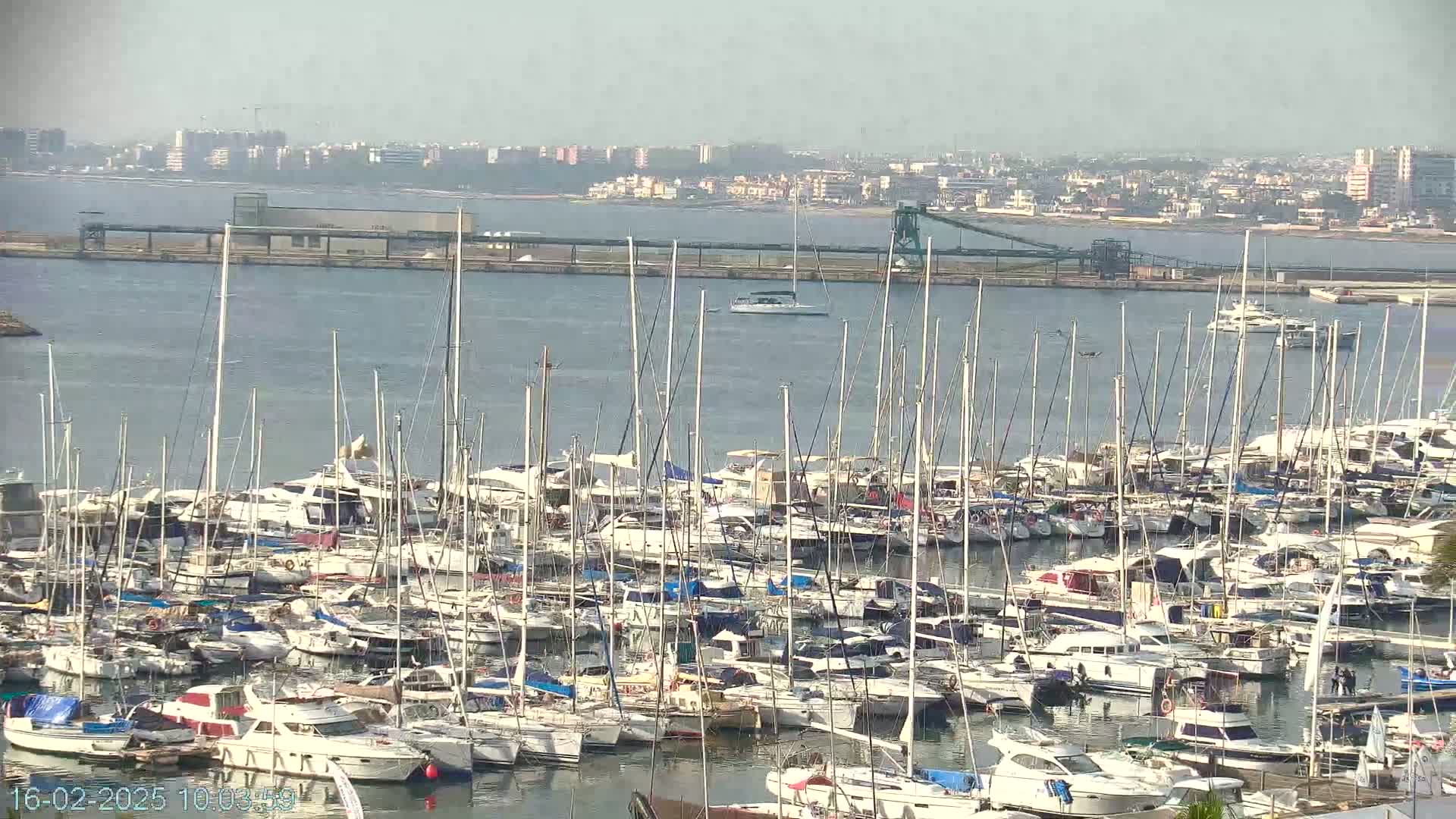 A large marina filled with numerous sailboats and motorboats is situated on calm water, with a distant cityscape visible under hazy skies.