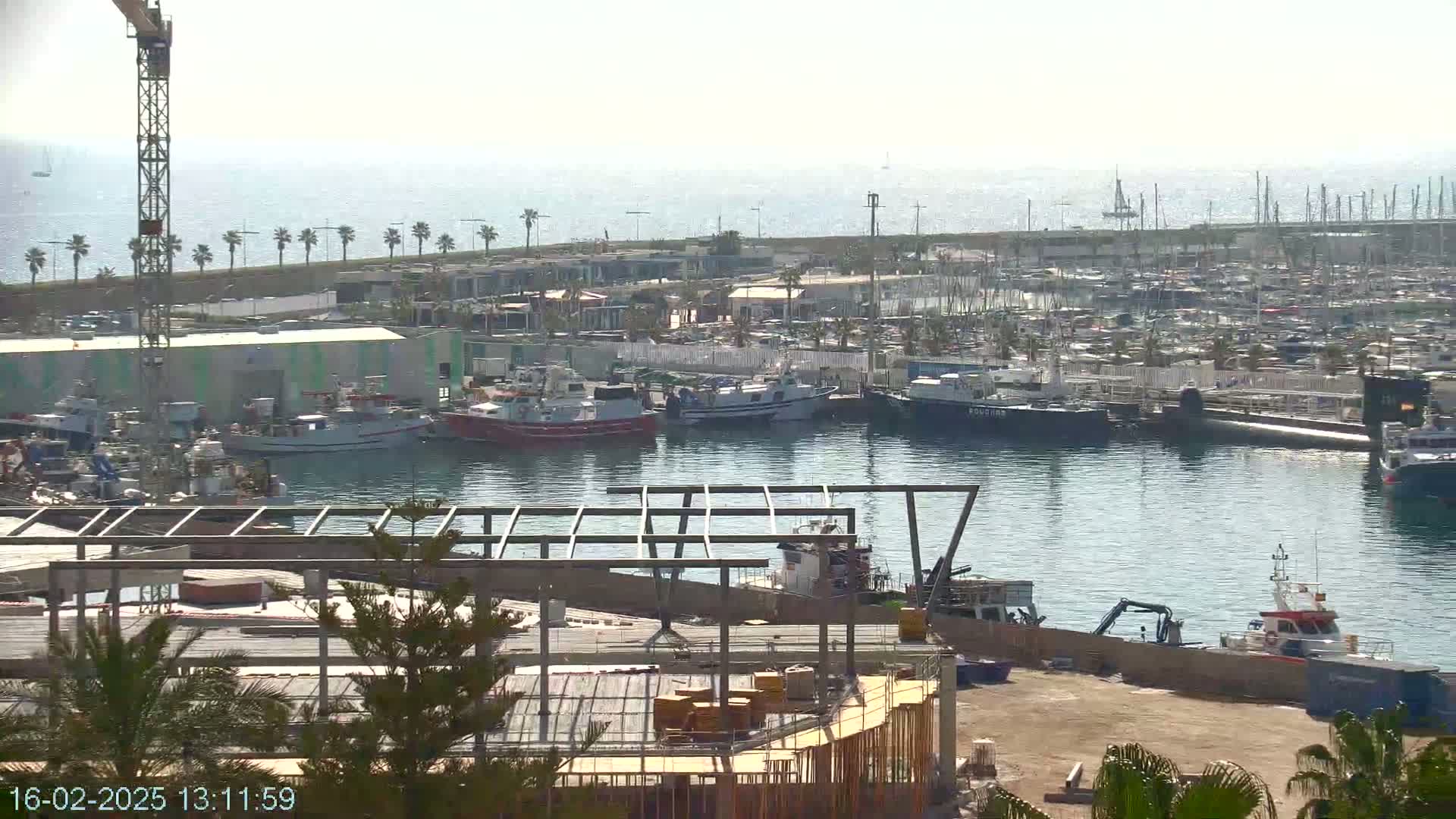 A harbor filled with numerous boats and ships under a clear, sunny sky, with a construction site in the foreground.