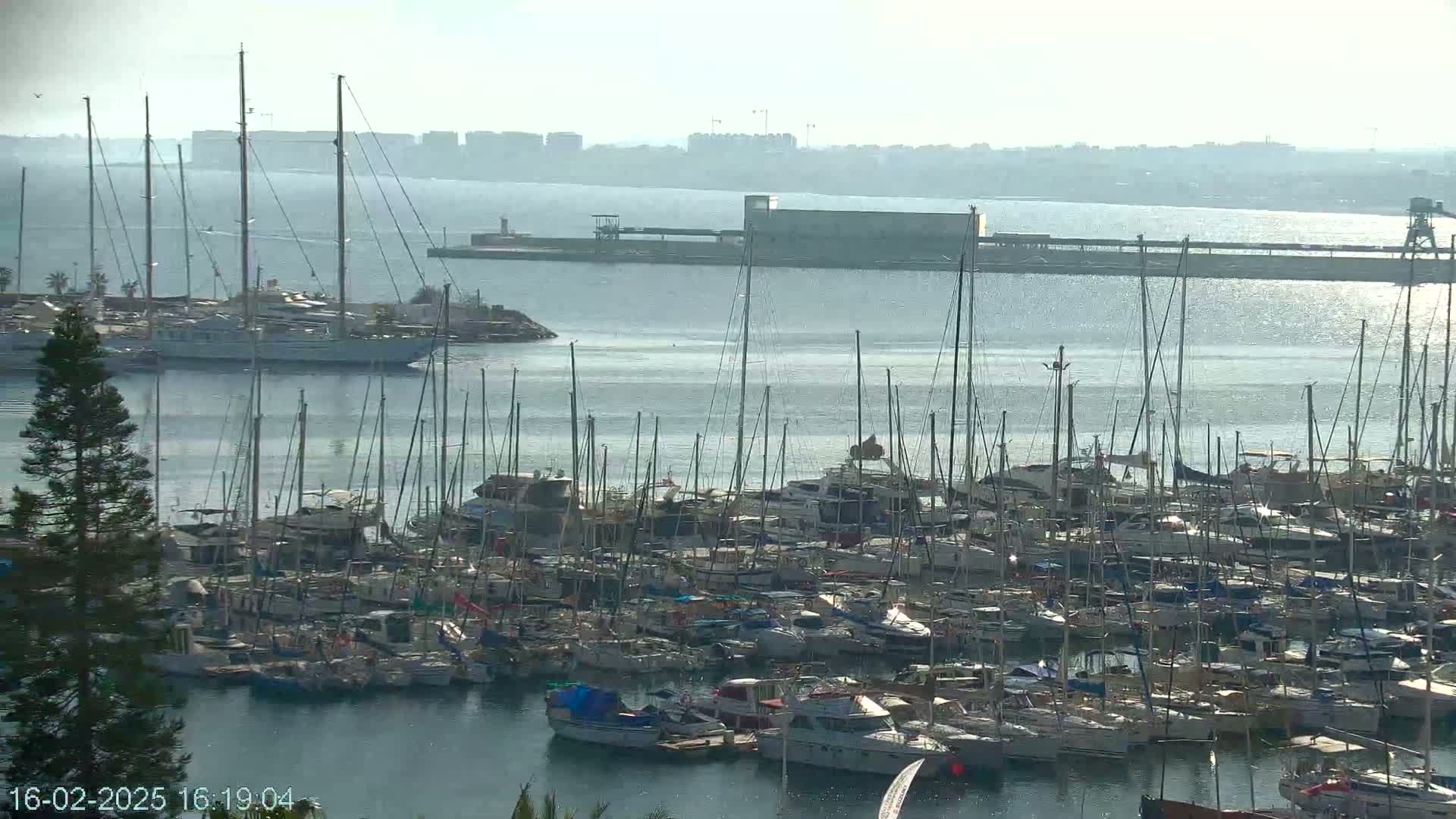 A large marina filled with numerous sailboats and motorboats is visible on a hazy day, with a larger vessel and distant cityscape in the background.
