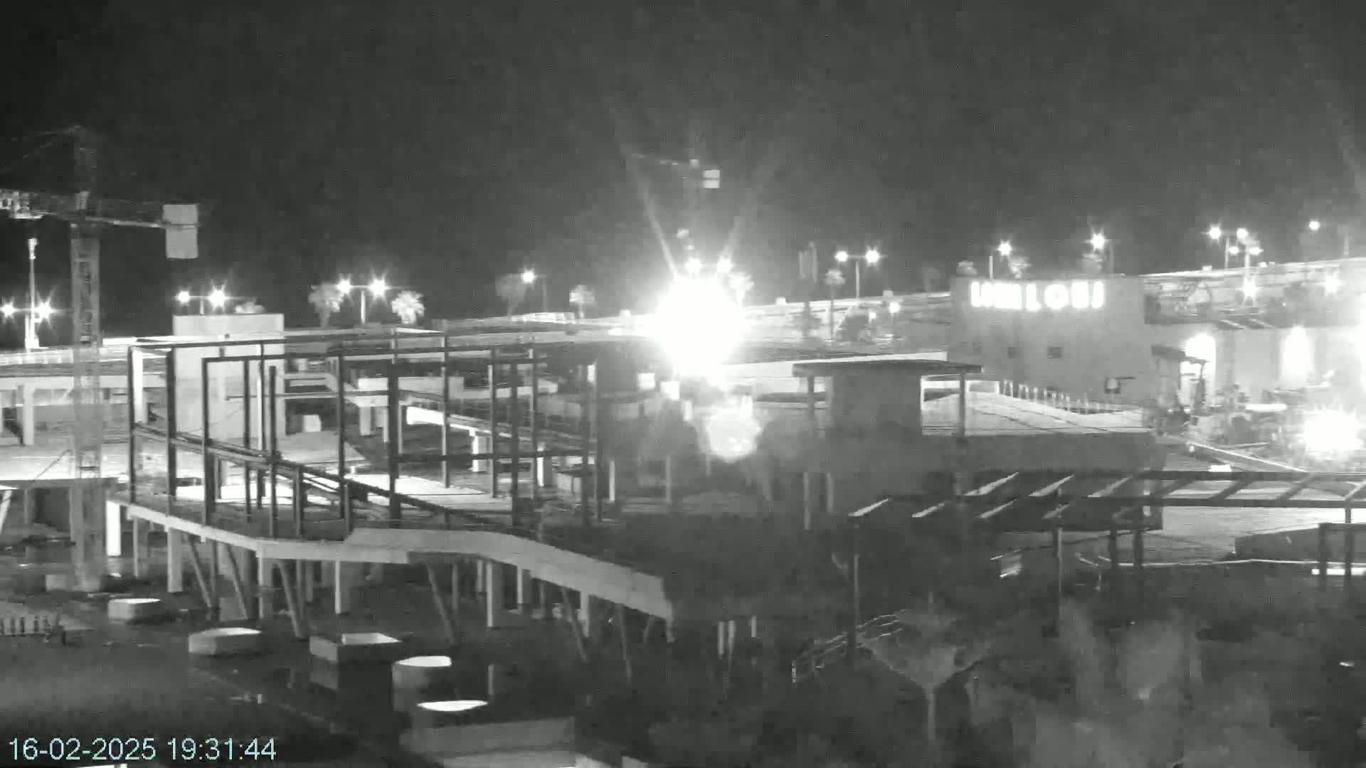 A nighttime, brightly lit construction site with a crane and skeletal framework of a building under construction next to an illuminated building.