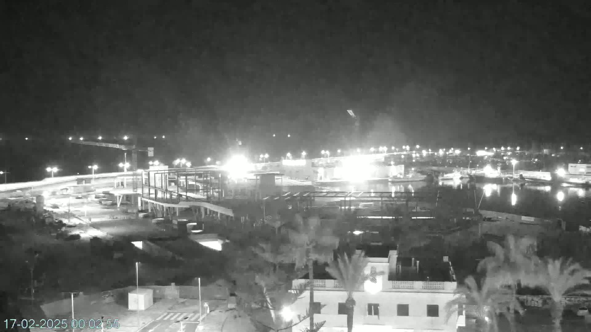 A nighttime, black and white view of a waterfront area with construction underway, illuminated by artificial lights.
