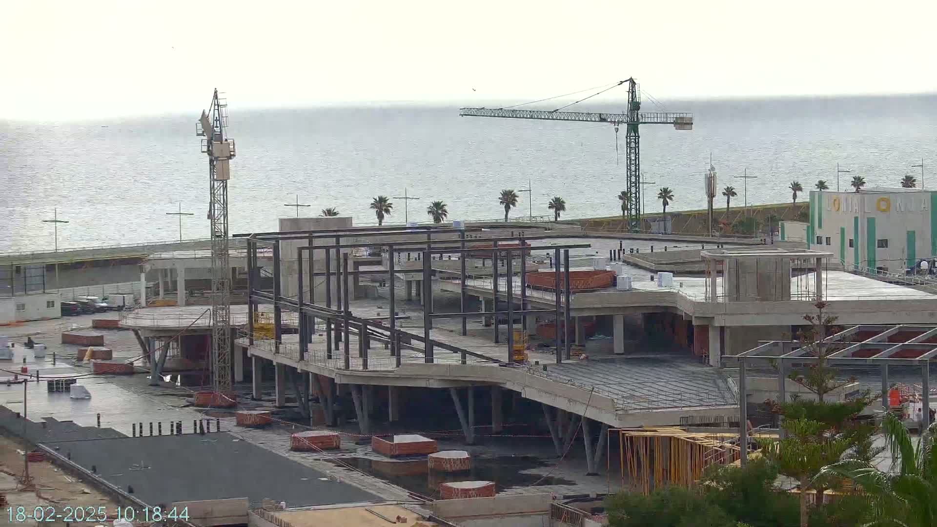A large building under construction near a body of water on an overcast day, with two construction cranes visible.