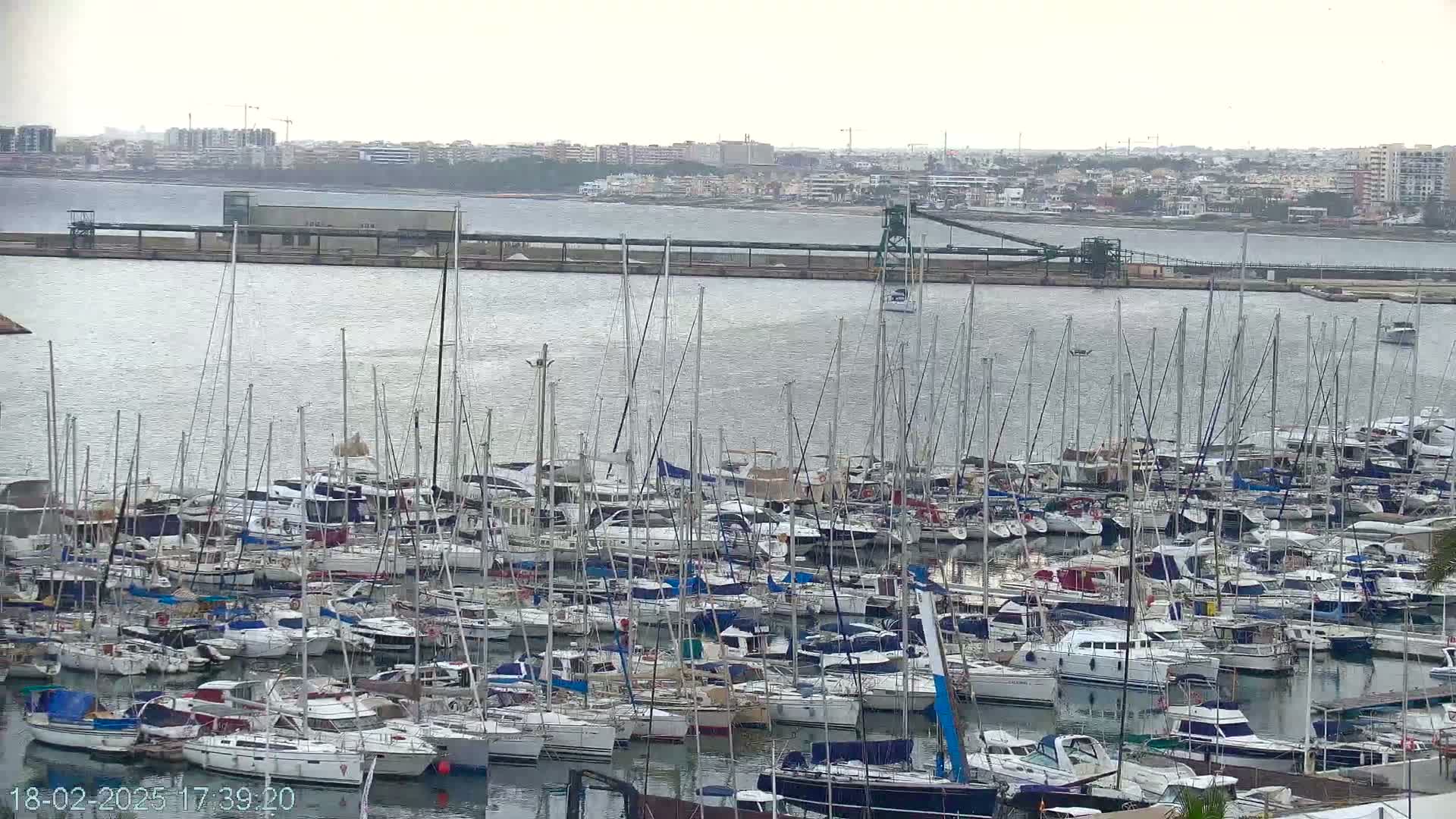 A large marina filled with numerous sailboats and motorboats is visible under an overcast sky.