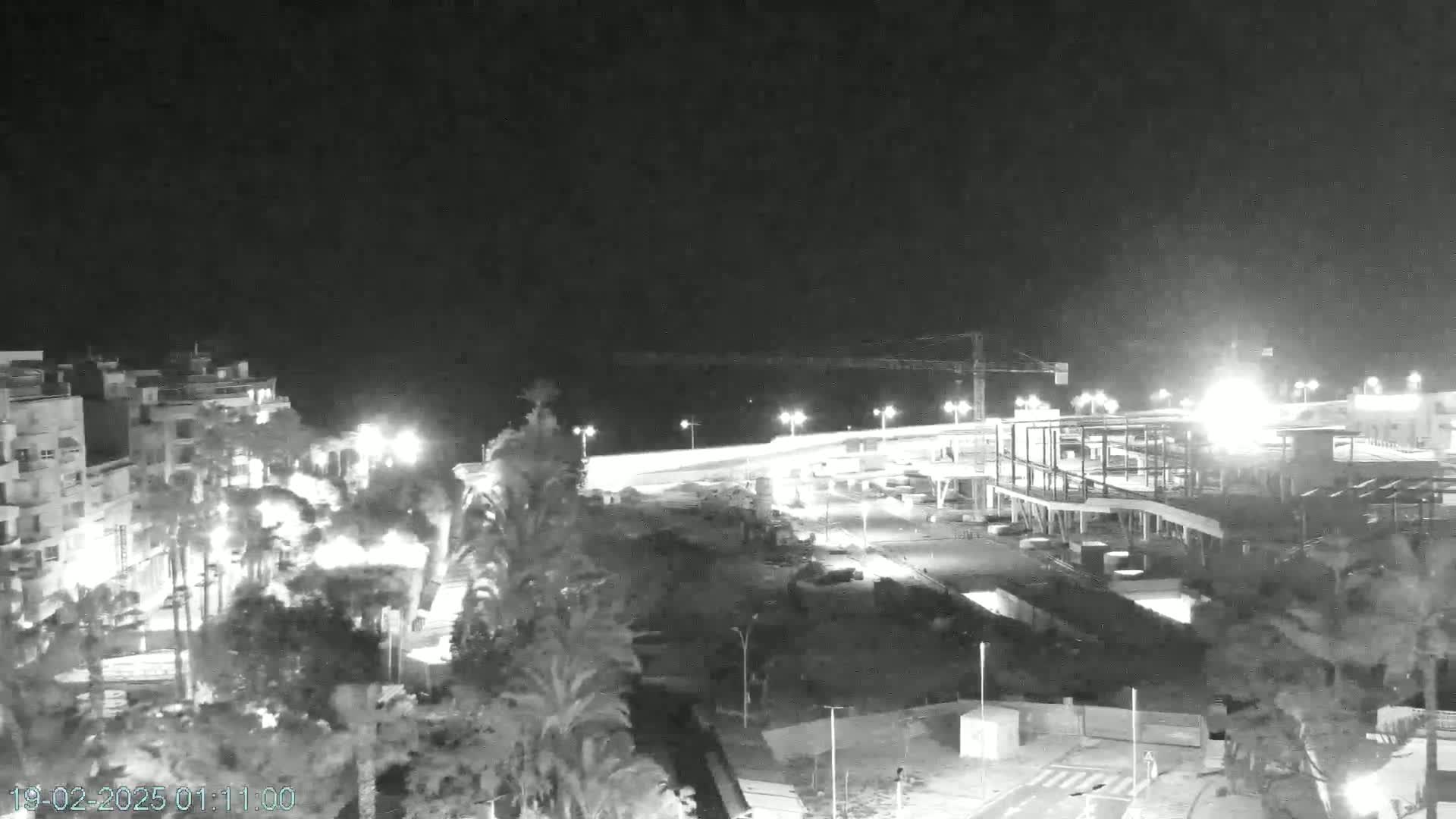 A nighttime, low-light view of a coastal town shows buildings under construction near a waterfront, illuminated by streetlights.