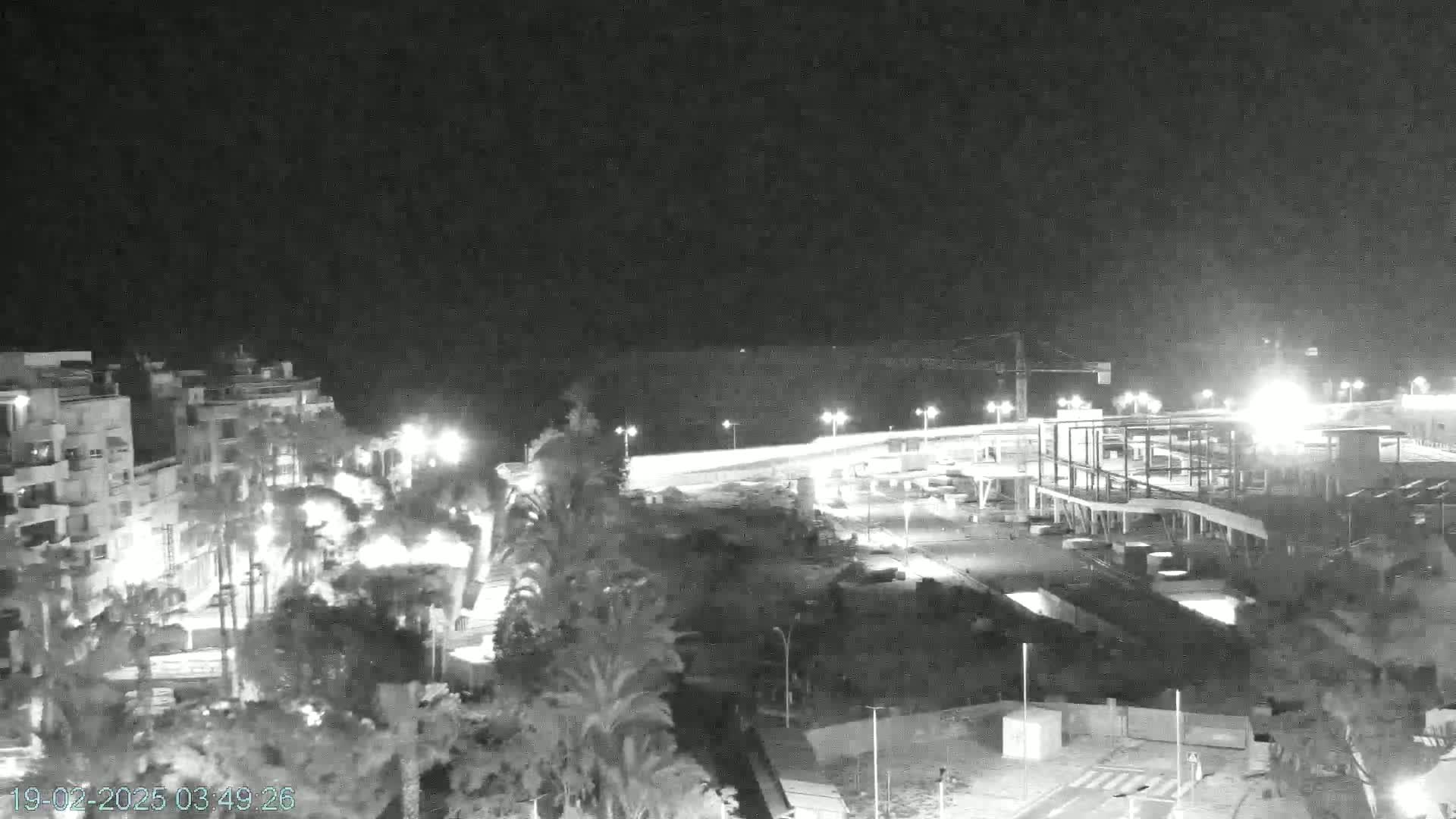 A nighttime, black and white view shows a coastal town with buildings, palm trees, and a partially-constructed structure near a brightly lit roadway.