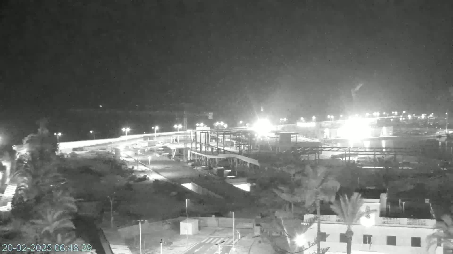 A nighttime, black and white view of a coastal area shows a partially constructed building near palm trees and brightly lit roadways.