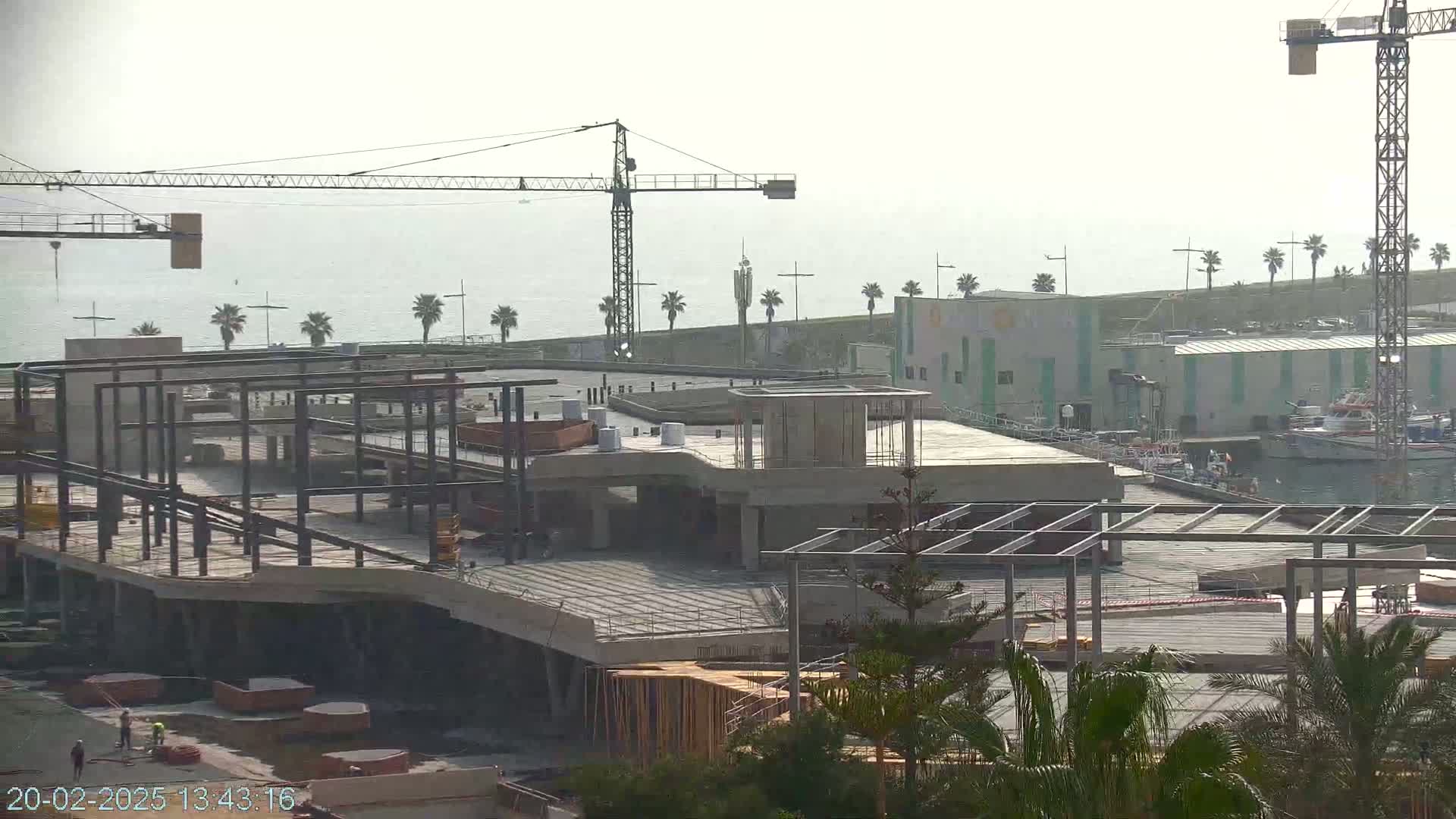 A partially constructed building with steel framework and concrete is situated near a calm body of water with palm trees and boats under a hazy sky.