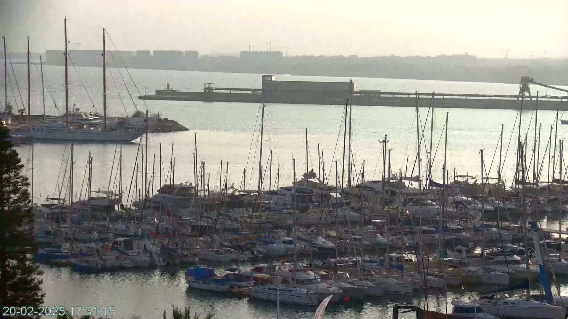 A marina filled with numerous sailboats and yachts is visible on a hazy day, with a large cargo ship and distant industrial structures in the background.