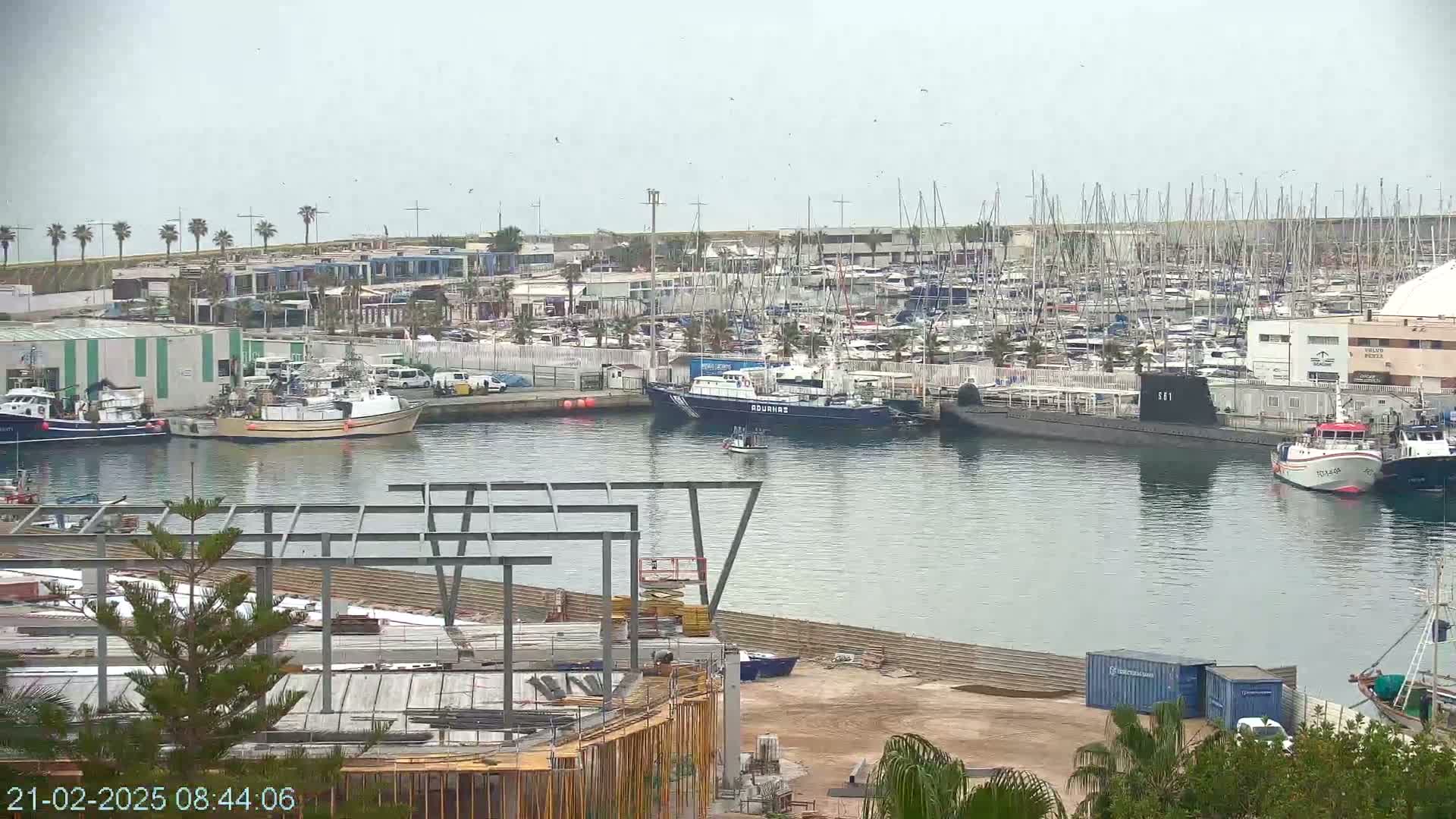 A harbor filled with numerous boats and ships, including a submarine, under an overcast sky.