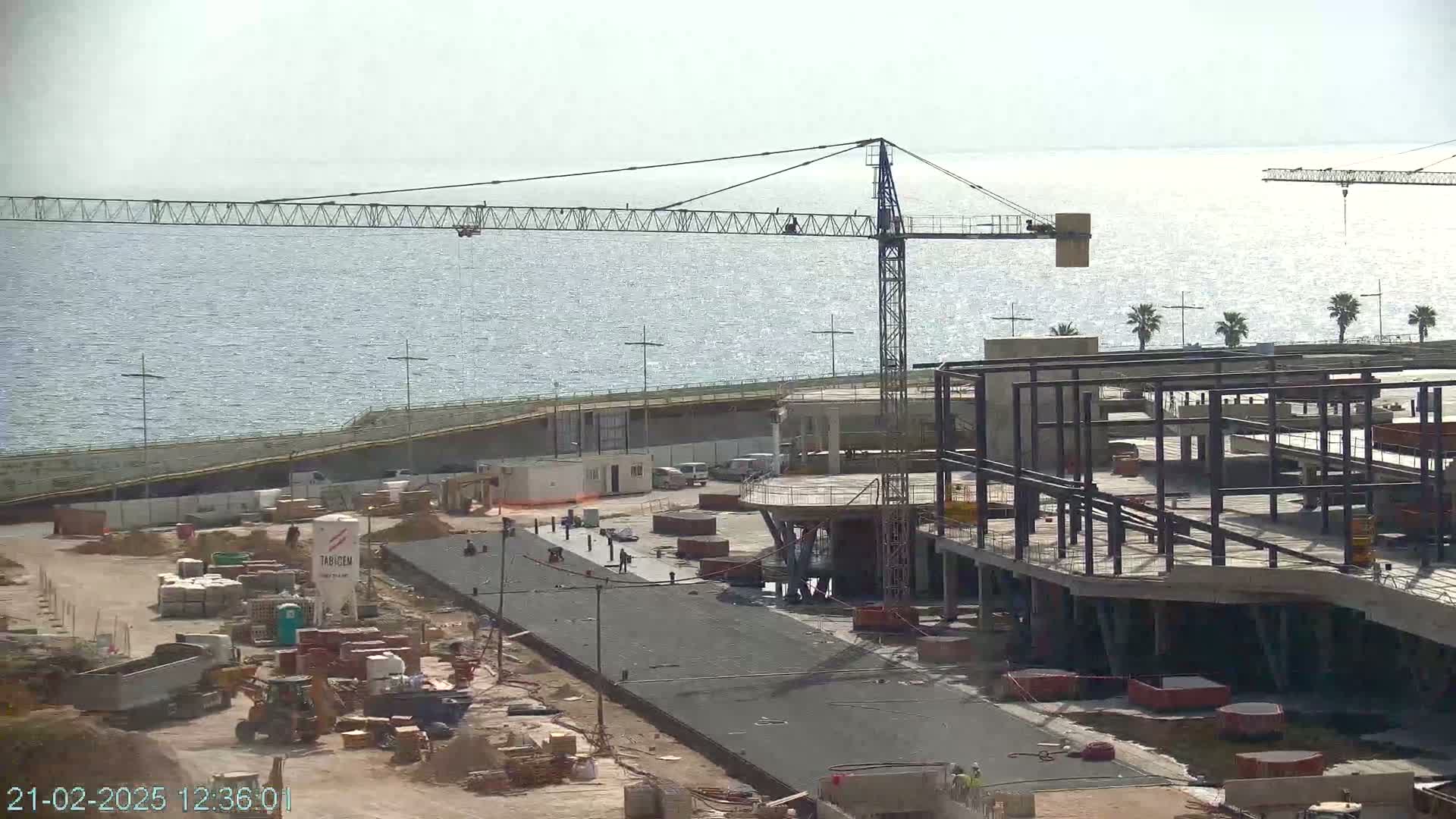 A construction site near a body of water under a mostly clear sky shows a large crane and the steel framework of a building under construction.