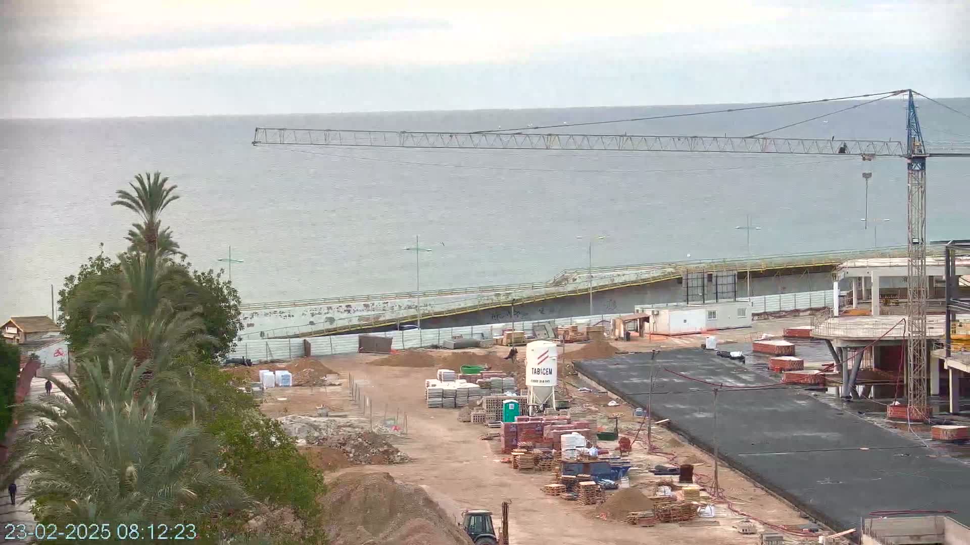 A construction site near a body of water under an overcast sky shows a crane, various building materials, and partially constructed buildings.