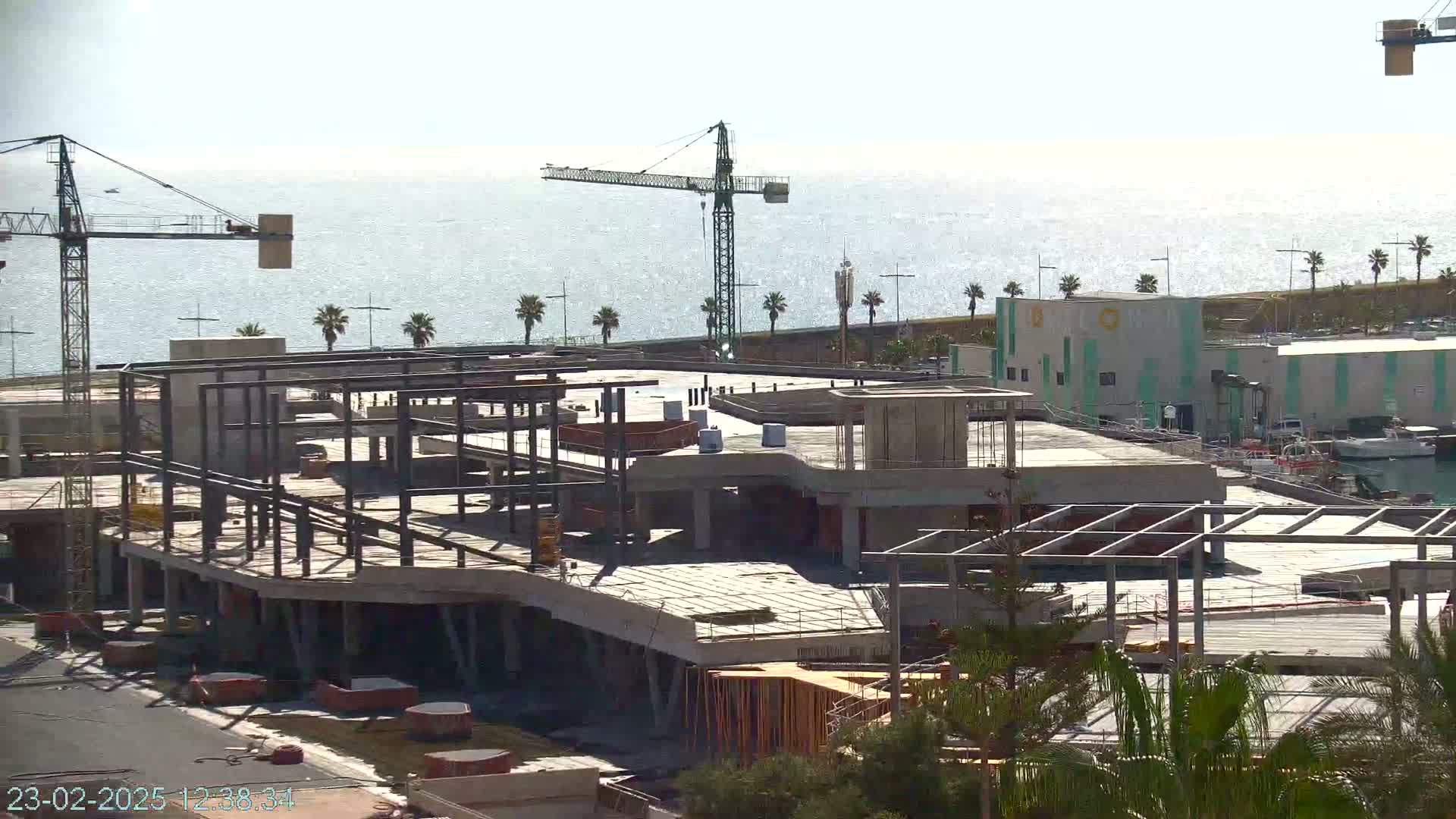 A large concrete structure under construction near a body of water on a sunny day, with several cranes visible in the background.