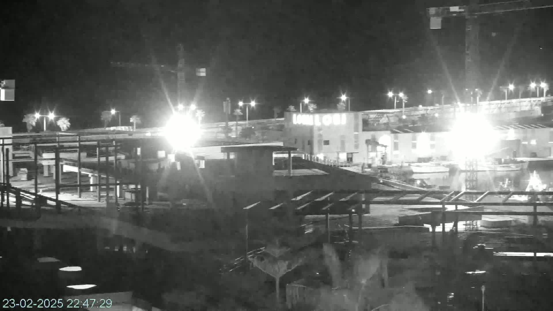 A nighttime, low-light image shows an area under construction, with bright lights illuminating various structures and ongoing work, some of which appears to be a building with a partially completed roof.