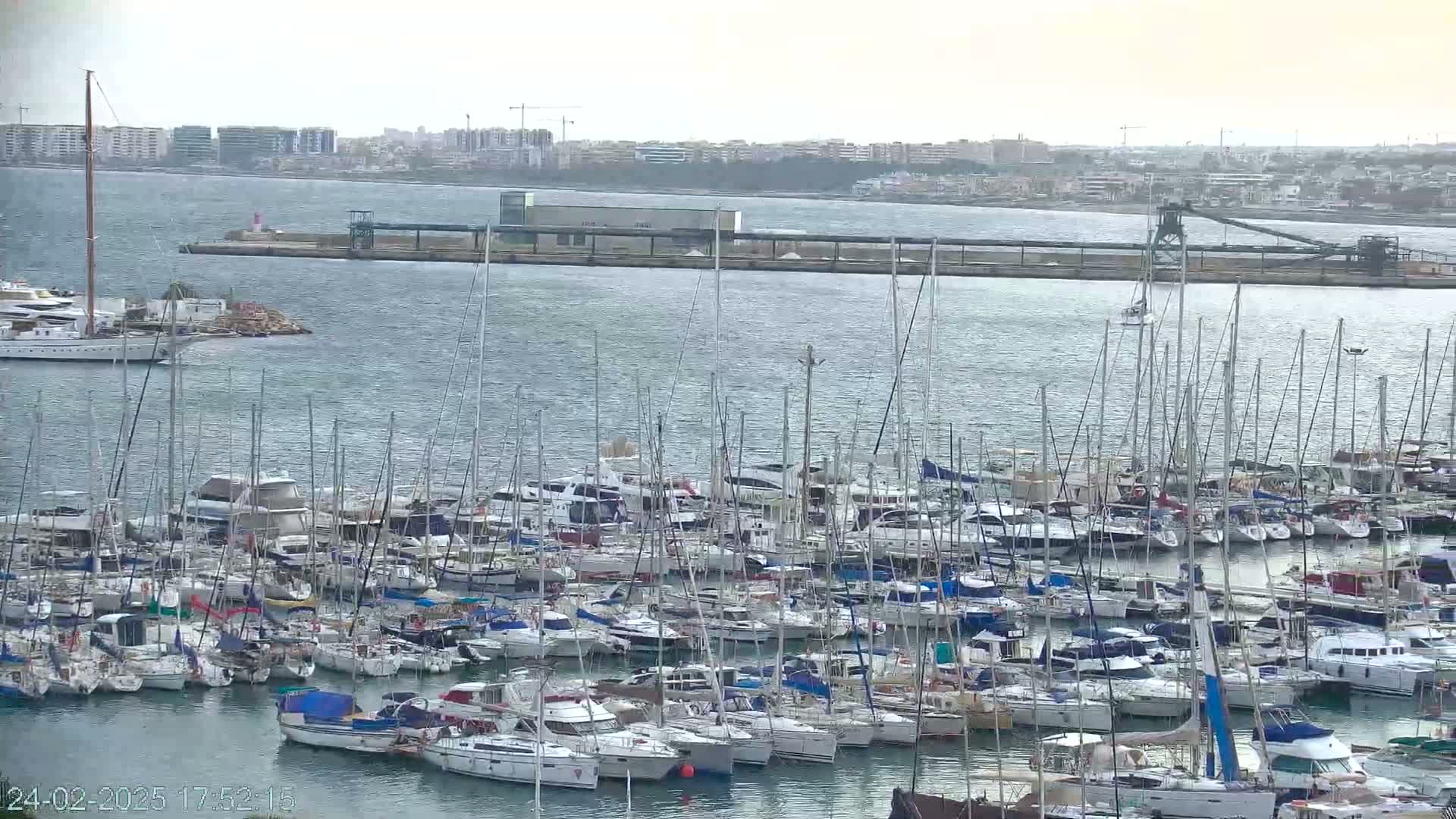 A large marina filled with numerous sailboats and motorboats is situated on calm water in front of a distant cityscape under an overcast sky.