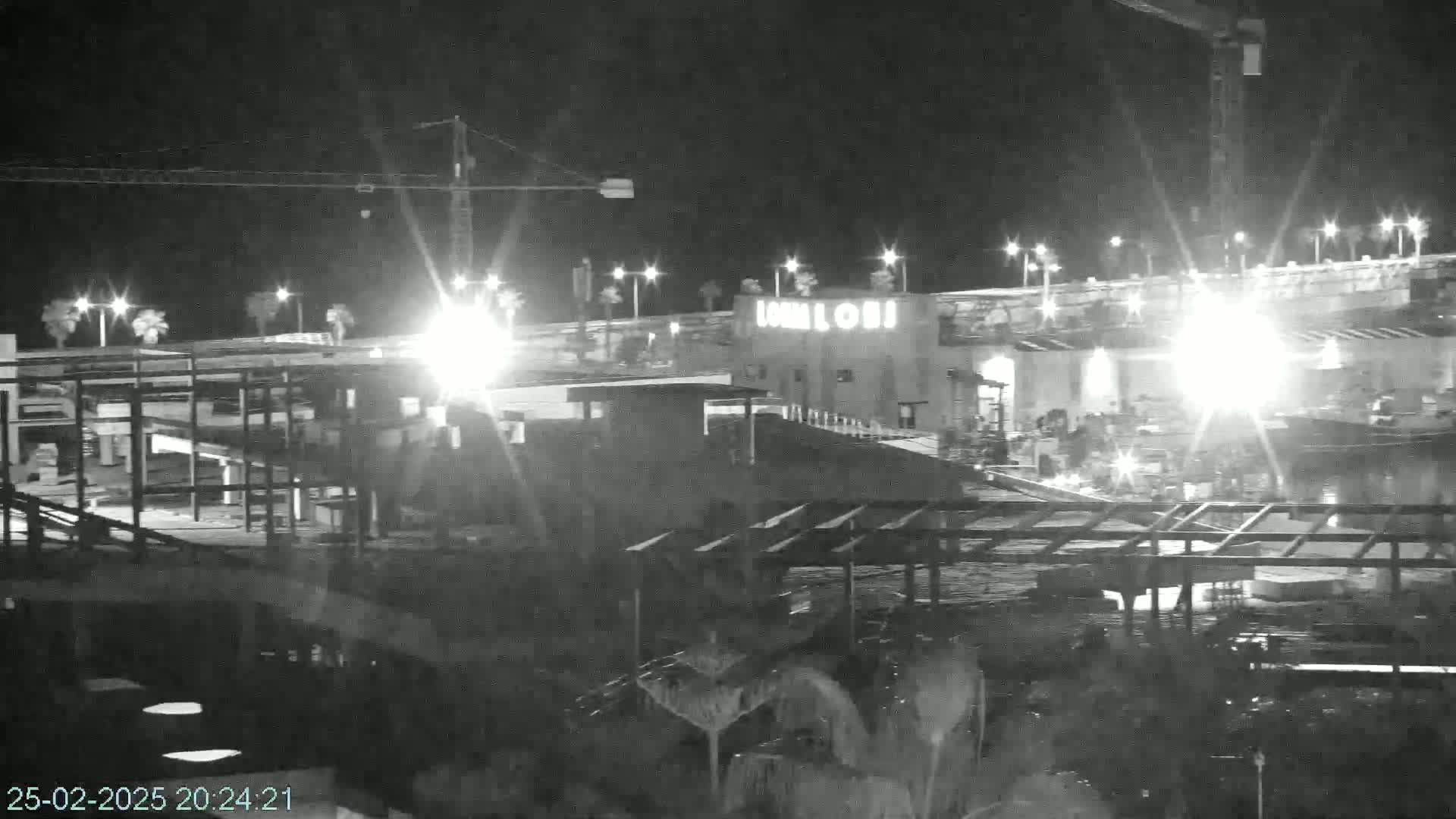 A nighttime, black-and-white view of a partly-constructed building complex under bright lights, with a crane visible in the background.