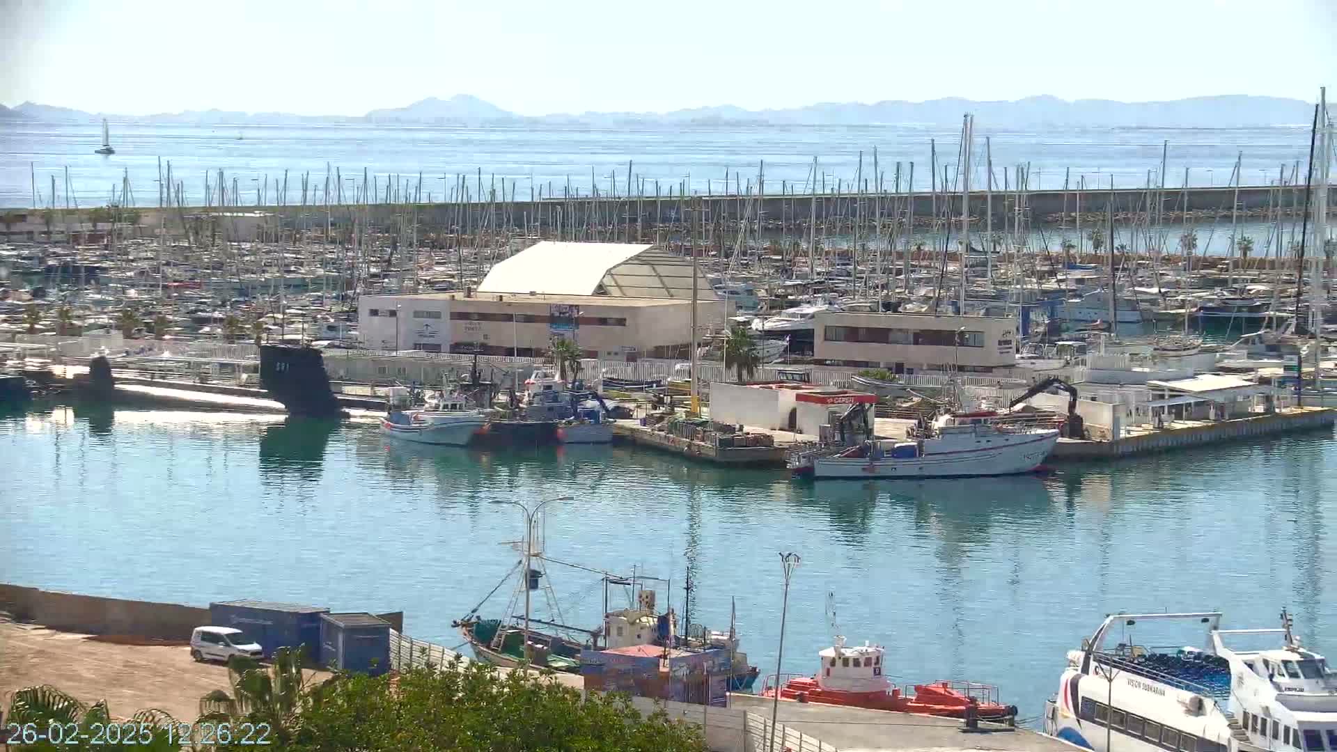 A sunny day shows a marina filled with sailboats and motorboats, a submarine, and several fishing vessels, with distant mountains visible in the background.
