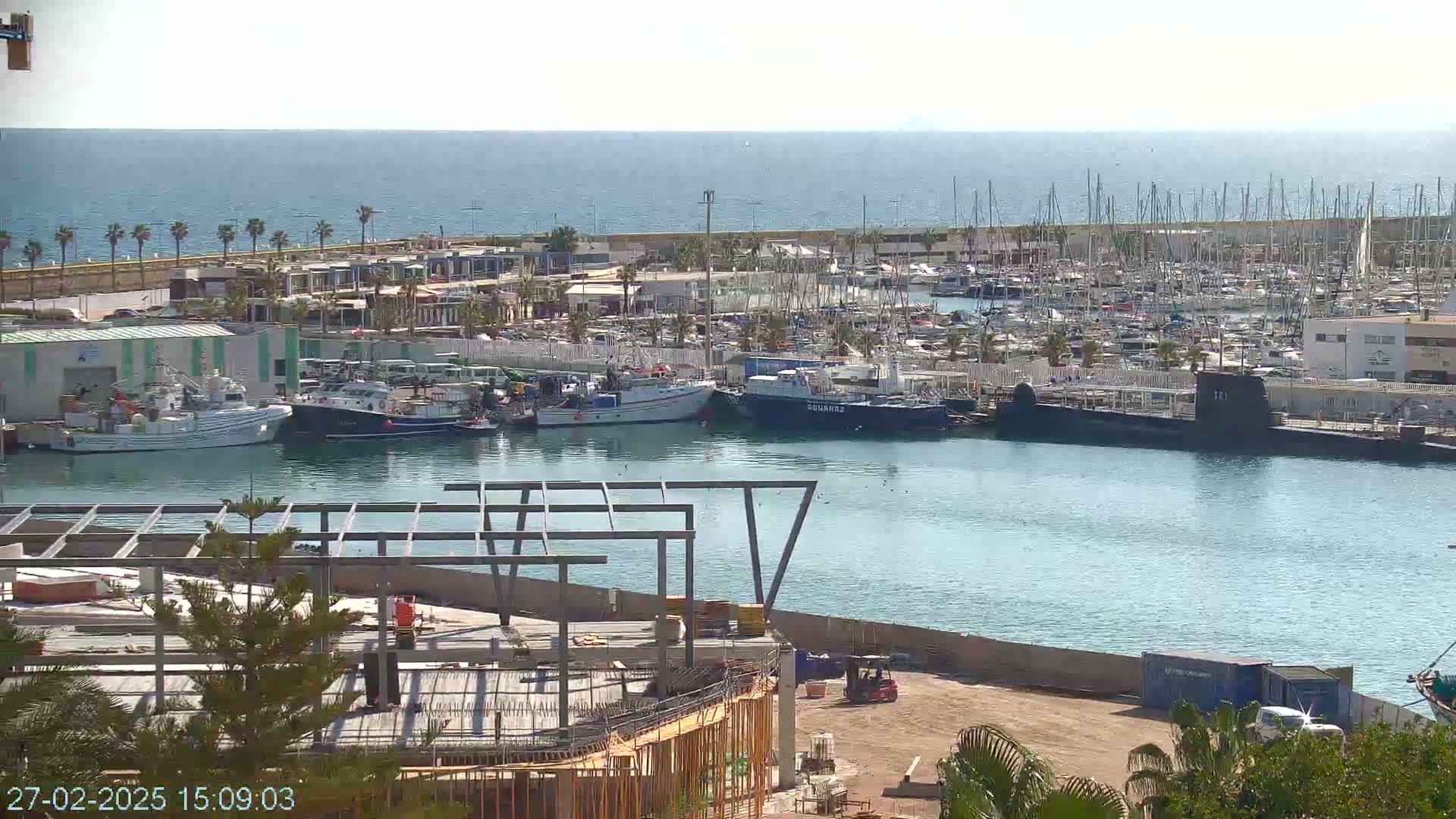A harbor filled with numerous boats and a submarine, under a sunny sky, is partially visible from an elevated vantage point overlooking a construction site.