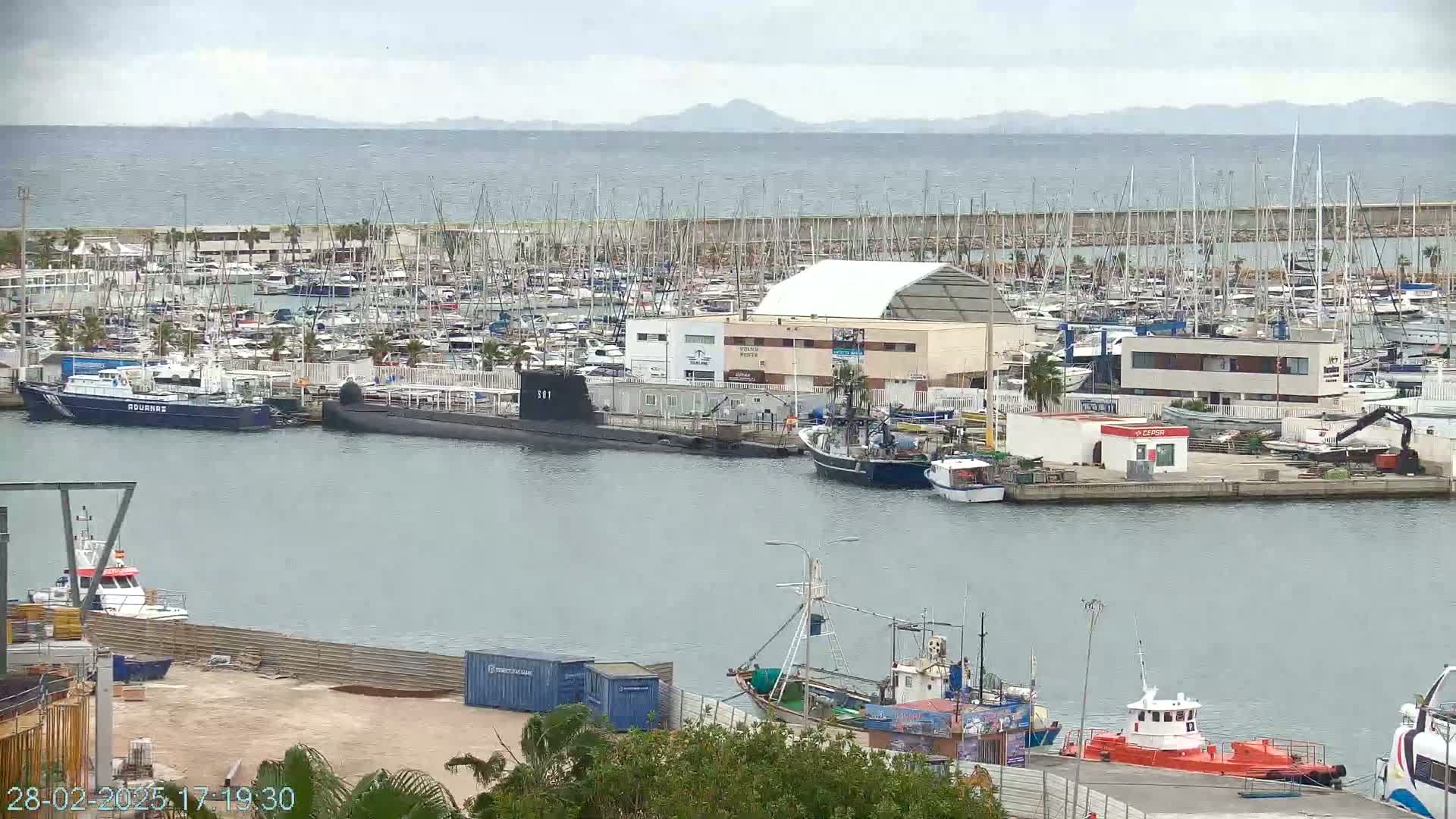 A harbor filled with numerous boats and a submarine, under an overcast sky, with a calm sea visible in the background.