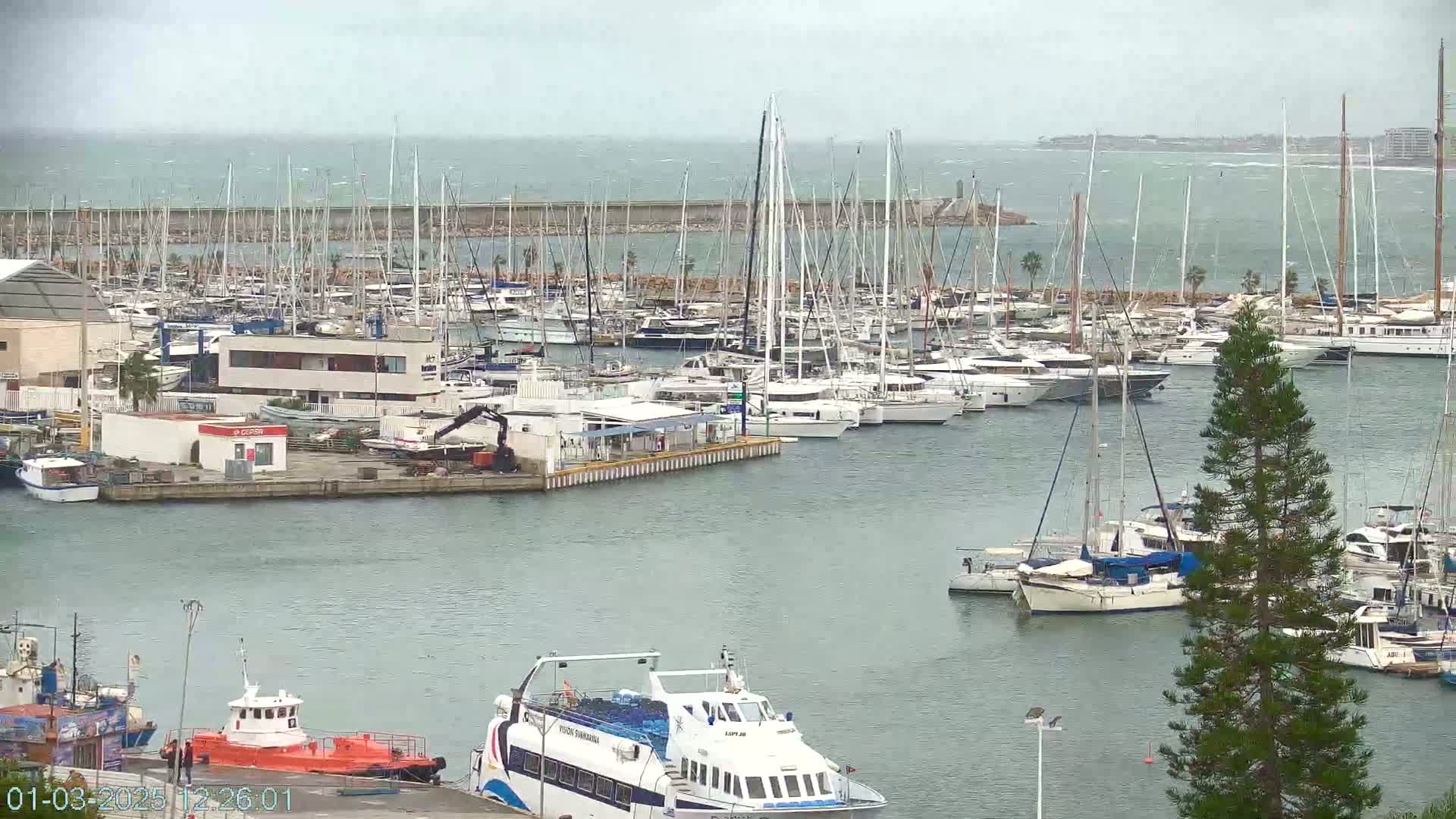 A marina filled with numerous sailboats and motorboats on a cloudy day with a choppy sea in the background.