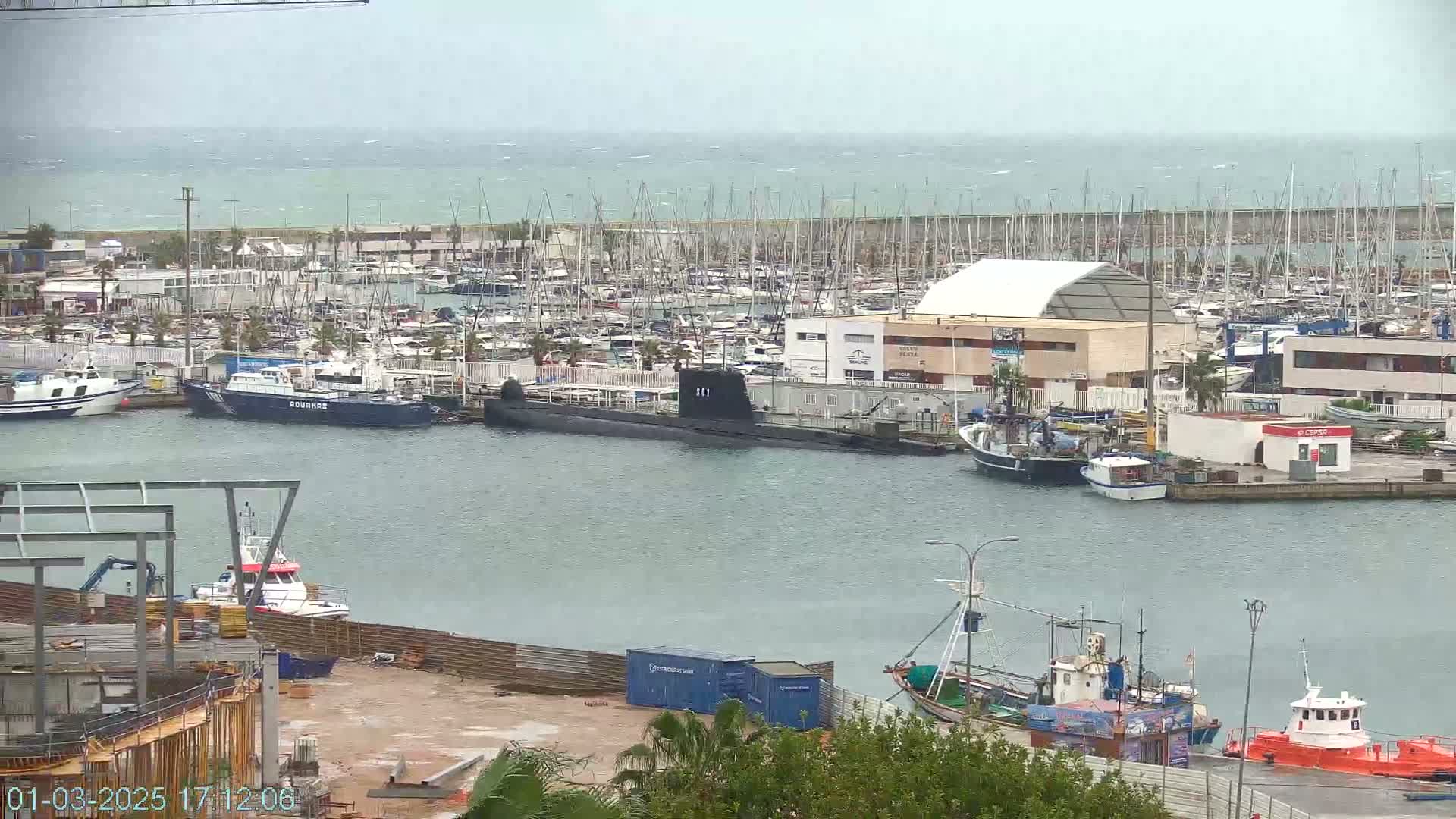 A harbor filled with numerous boats and a submarine, under an overcast sky with a choppy sea in the distance.
