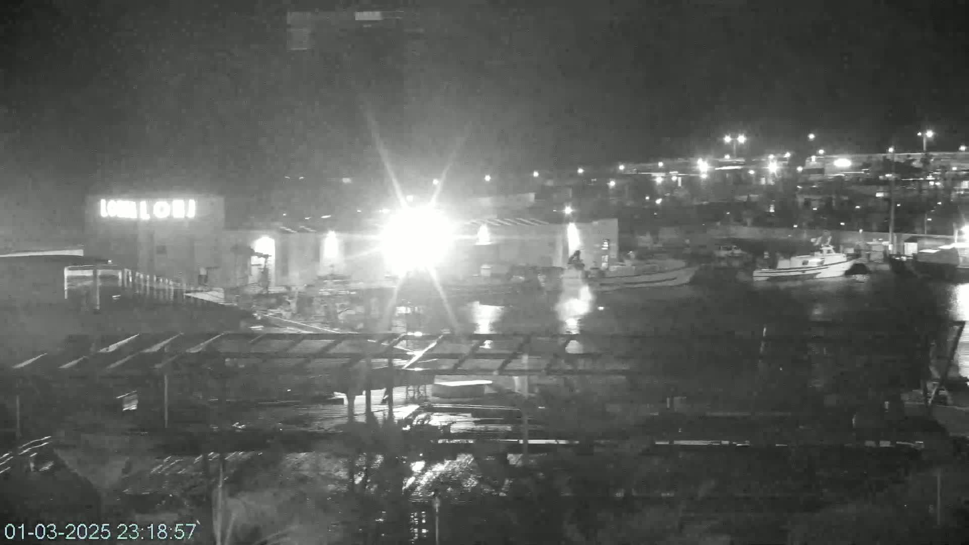 A nighttime, possibly rainy, view of a harbor with several boats moored near a building with lights.