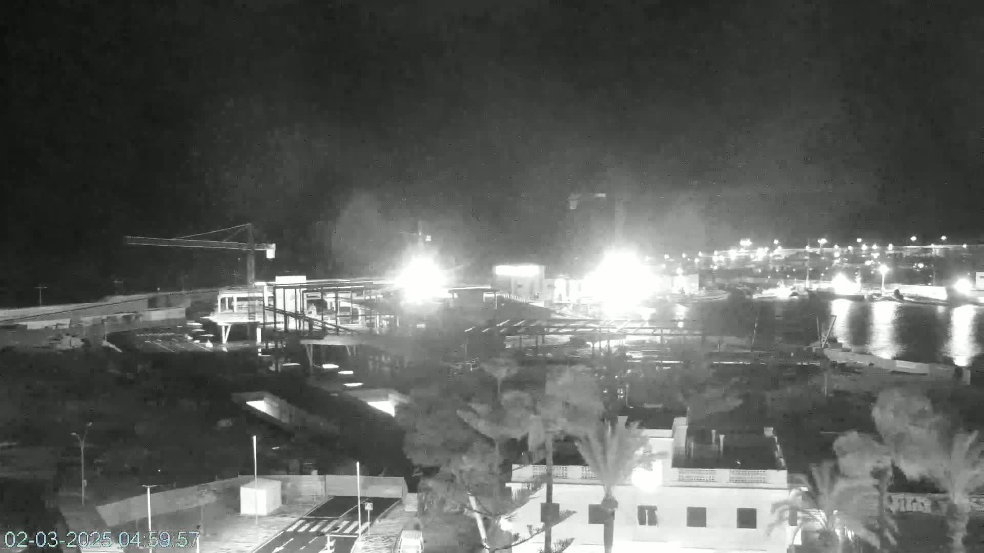 A nighttime, high-angle view of a harbor with construction underway near a building and palm trees under a dark sky.
