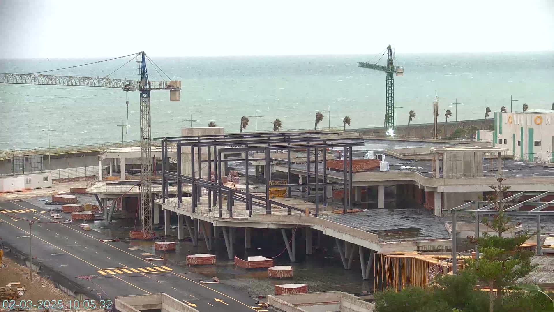 A large building under construction near a body of water on a windy, overcast day, with two construction cranes visible.