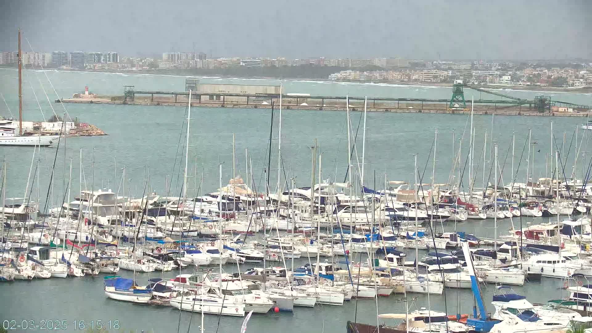 A marina filled with numerous sailboats and motorboats is visible under an overcast sky.