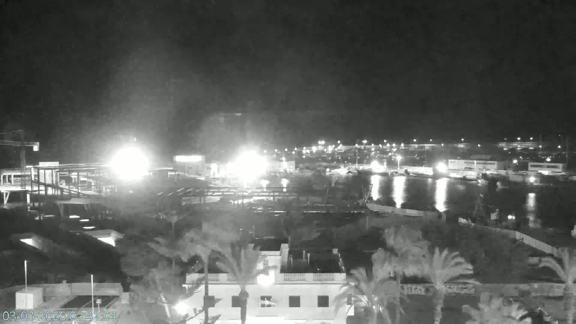 A nighttime, low-light view of a harbor with boats and buildings, including a partially constructed structure in the foreground, under a clear sky.