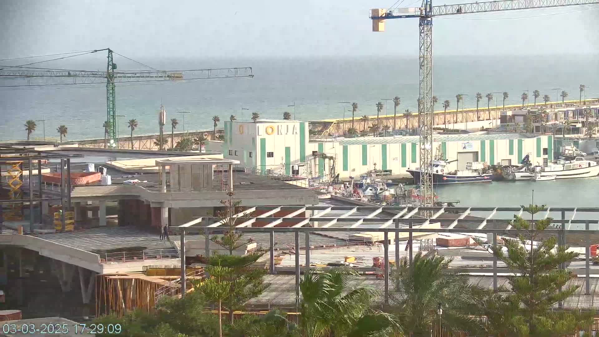 A construction site with a partially built structure is adjacent to a harbor with several boats docked, under a hazy sky.
