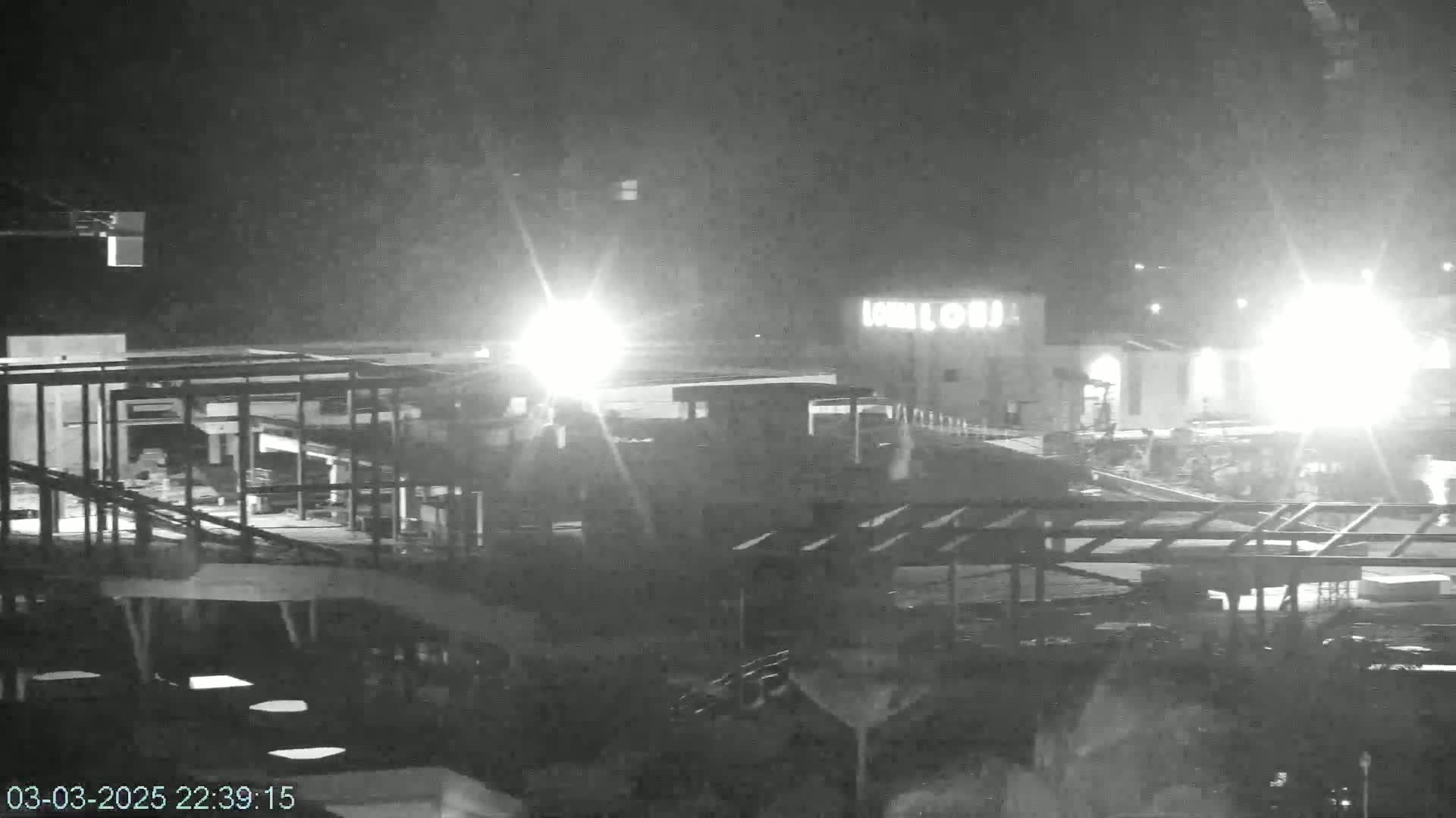 A nighttime, low-light view of an outdoor construction site with a partially built structure, illuminated by bright lights, next to a building.