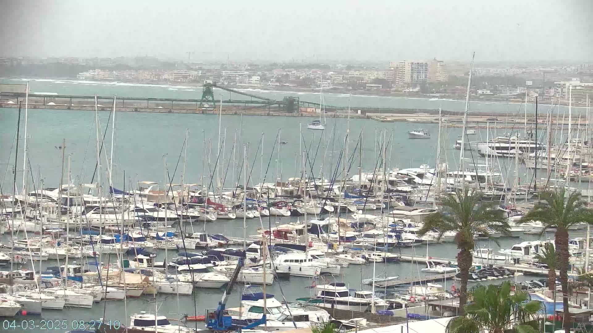 A hazy, overcast day reveals a densely packed marina filled with numerous sailboats and motorboats, situated near a city and industrial structures.
