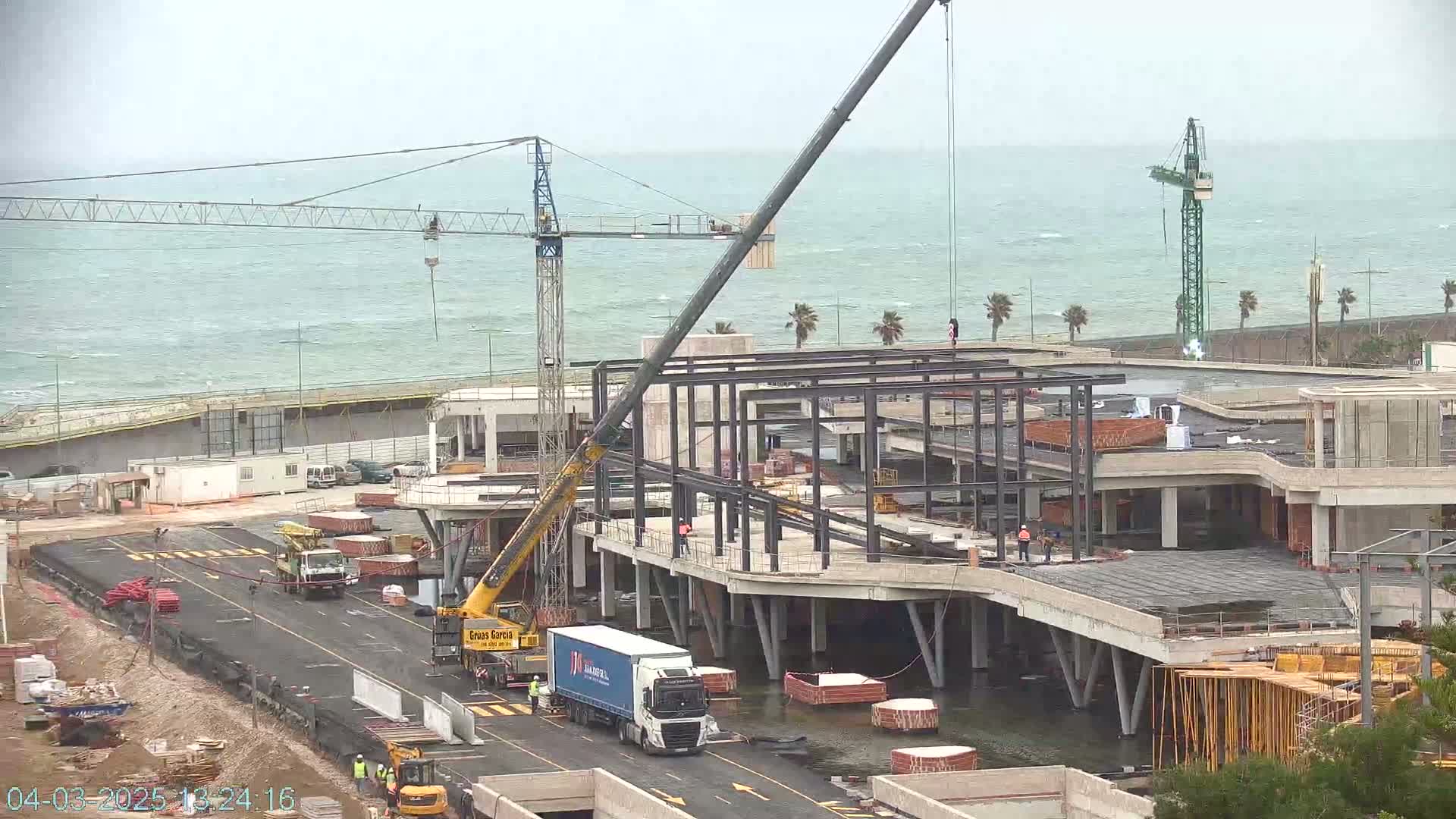 A large construction crane lifts a metal structure at a seaside construction site under partly cloudy skies and a choppy sea.