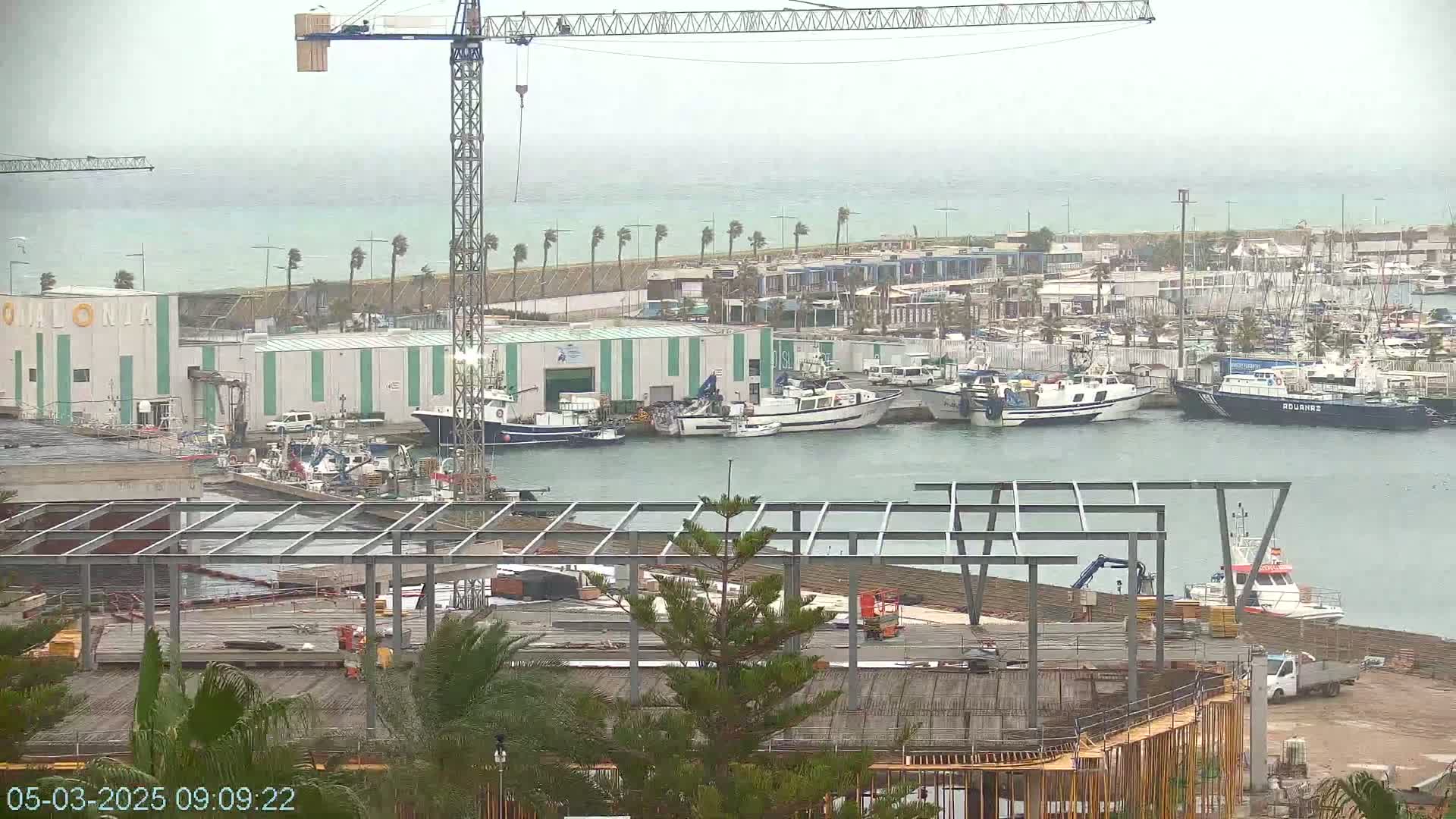 A harbor with numerous boats, a construction site, and a large crane is visible under an overcast sky.