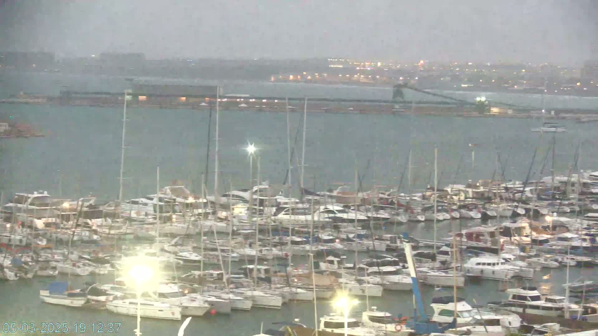 A marina filled with many boats is seen in the rain under an overcast sky.