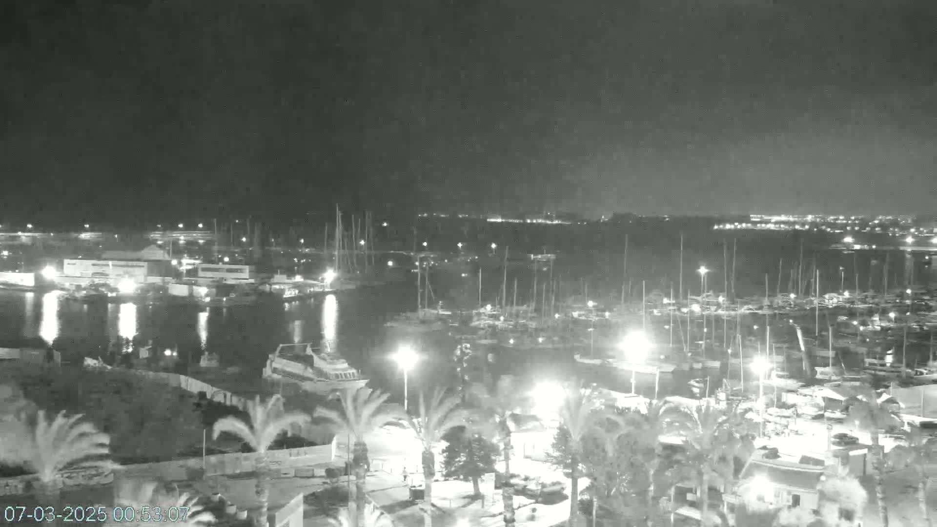 A nighttime view of a harbor filled with boats, illuminated by lights, under a dark, possibly overcast sky.