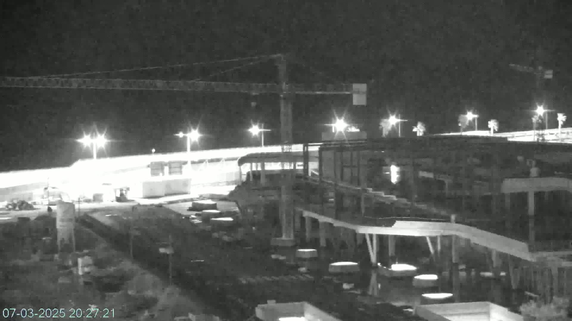 A nighttime, black and white image shows a construction site with a large building under construction, illuminated by artificial lights, with a construction crane visible in the background.