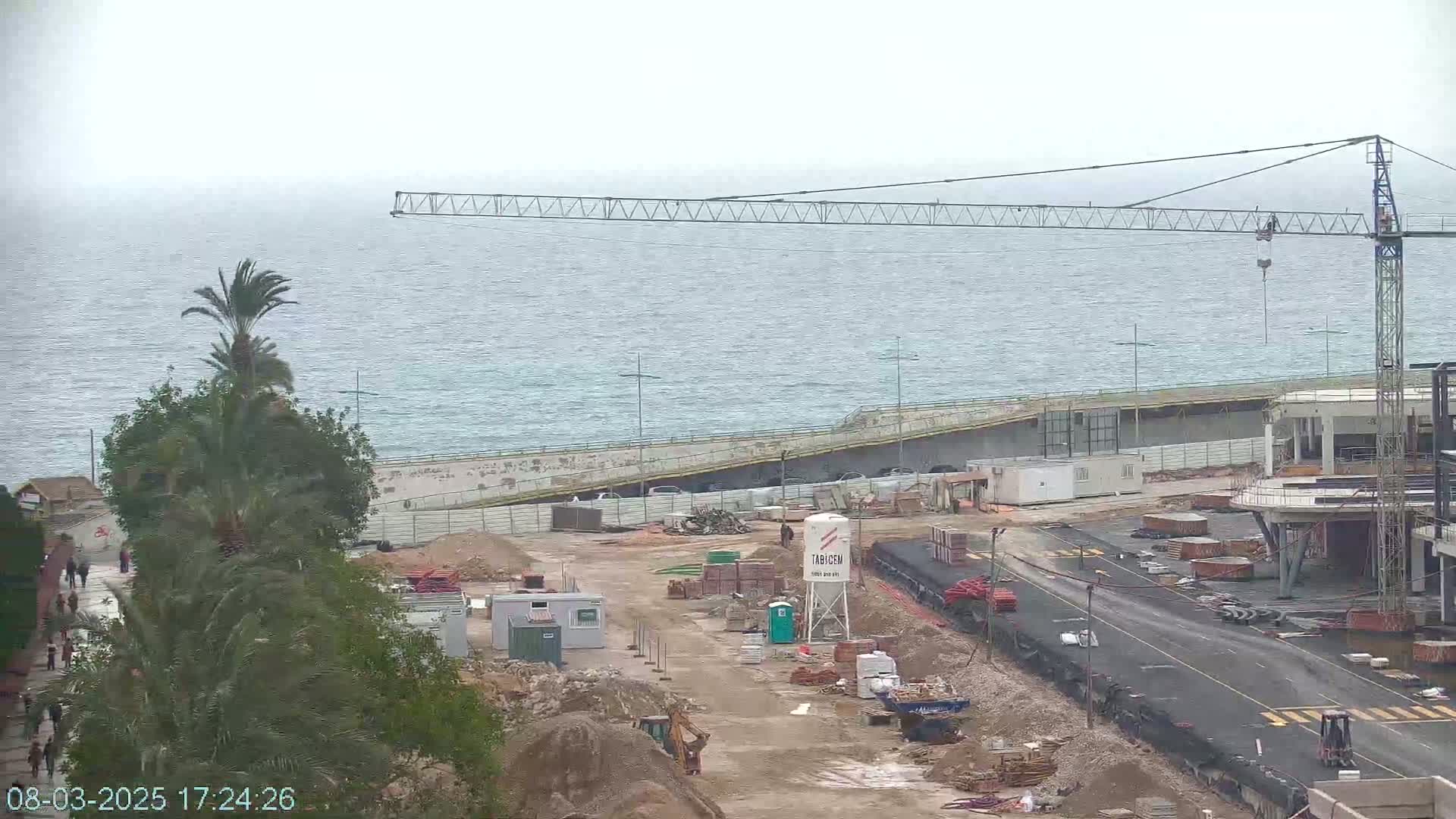 A construction site near a calm sea under an overcast sky, featuring a large crane and various building materials.
