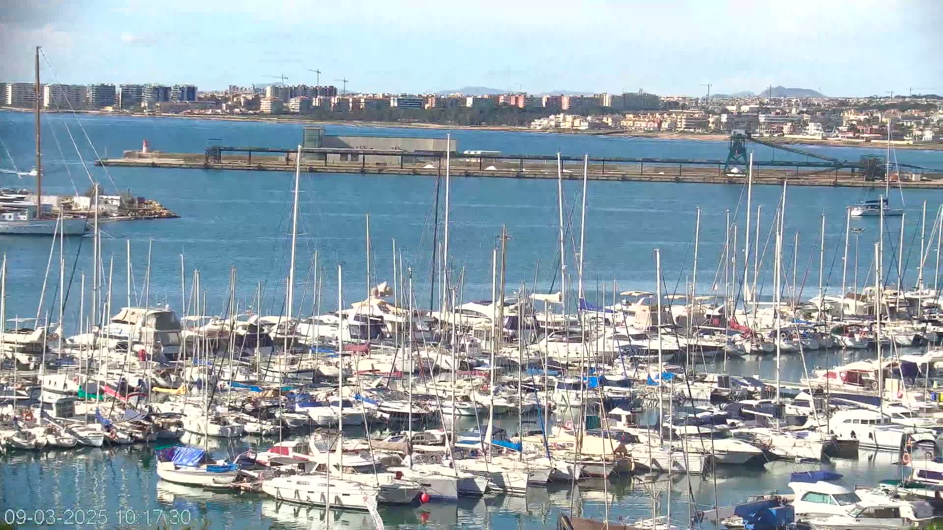 A large marina filled with numerous sailboats and motor yachts is situated on a calm body of water under a sunny sky, with a distant cityscape visible in the background.