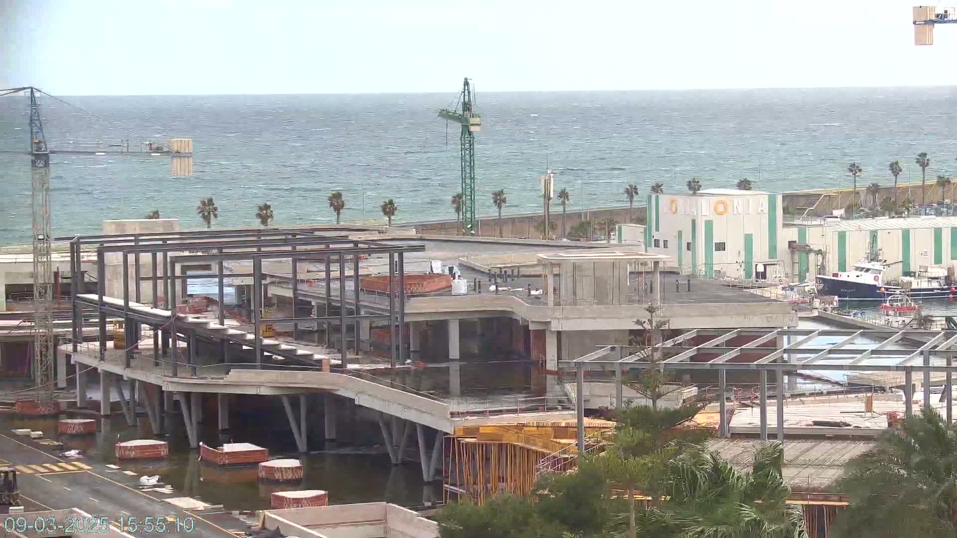 A partially constructed building near a body of water under a slightly overcast sky, with a crane and several boats visible in the distance.