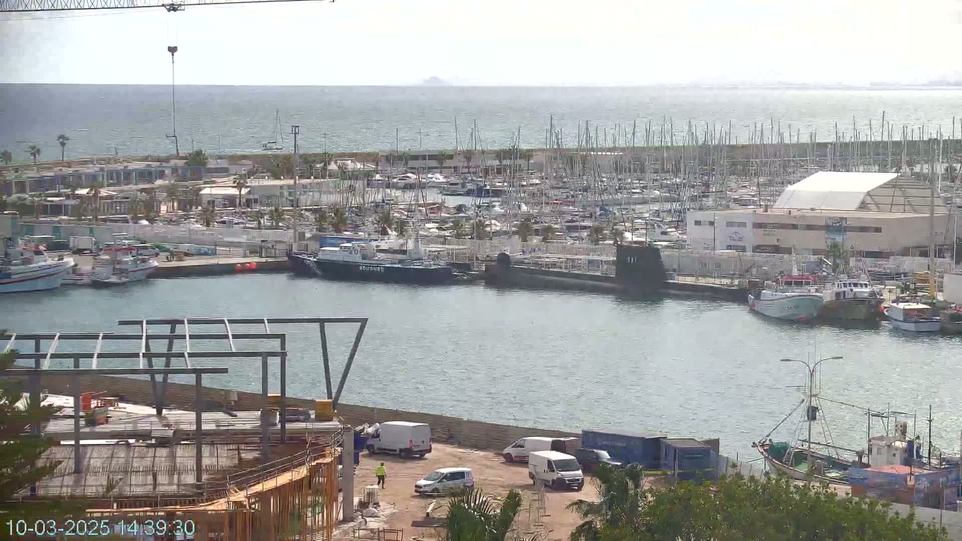 A harbor filled with numerous boats and a submarine, under a partly cloudy sky, with construction visible on the shore.