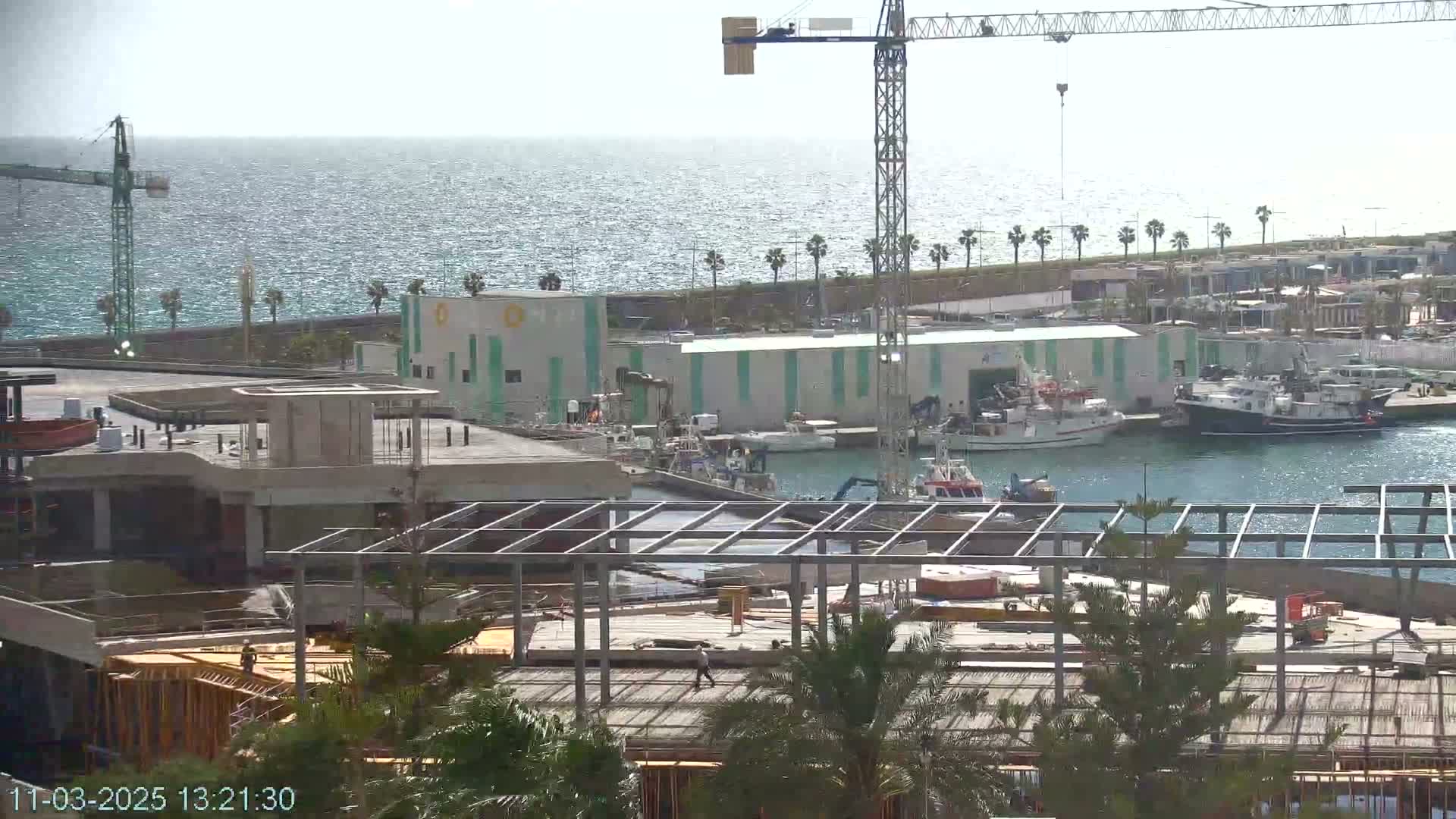 A partially constructed building near a harbor with several boats, a crane, and palm trees under a bright, sunny sky.