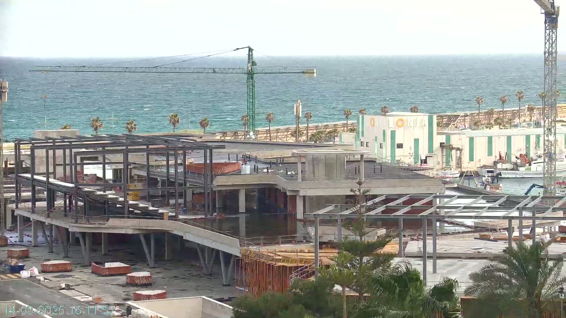 A partially constructed building with a steel framework sits near a coastline under a partly cloudy sky, with a crane visible in the background.